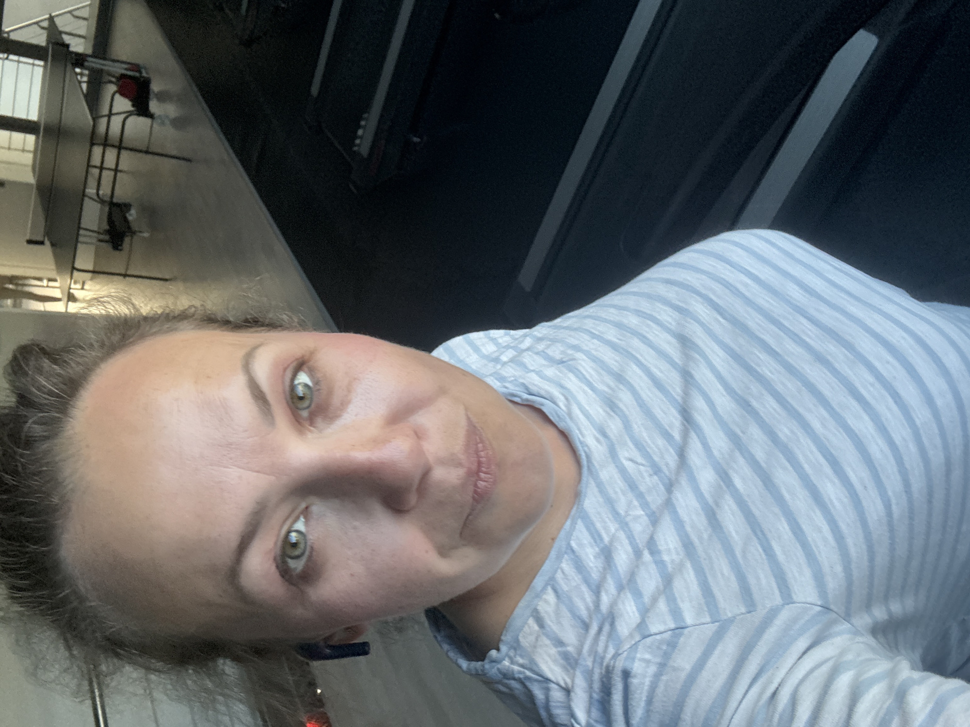 A woman with blonde hair pulled back is taking a selfie in a gym. She is wearing a light blue and white striped t-shirt and has earbuds in her ears. Her eyes are green and she is looking directly at the camera with a slight smile. In the background, there are treadmills and a ping pong table. The floor is light-colored and reflective.
