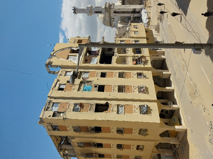 The image displays an urban street scene in Khan Yunis, Palestinian Territory, marked by extensive structural damage. A multi-story residential or commercial building, approximately six to seven floors high, occupies the left and central foreground. Its light tan facade is riddled with numerous impact holes, and many windows are shattered or missing, with torn fabric and debris visible within openings. The upper floors exhibit partial structural collapse and damage to balconies. The ground floor features arched supports, under which a yellow vehicle, a silver sedan, and another light-colored vehicle are parked.
In the background, to the right, stands a white mosque with a tall, slender minaret topped by a domed section, alongside a larger central dome. This mosque also shows significant structural damage, with visible crumbling and apertures in its walls and base.
The street below is a paved road, partially obscured by sand and scattered rubble, with remnants of white lane markings visible. A grey utility pole, equipped with horizontal power lines, stands prominently in the mid-ground on the right side of the street. Several individuals, appearing to be pedestrians, walk on the street in the mid-ground and background. The scene is observed under daylight conditions with a clear blue sky interspersed with white clouds.