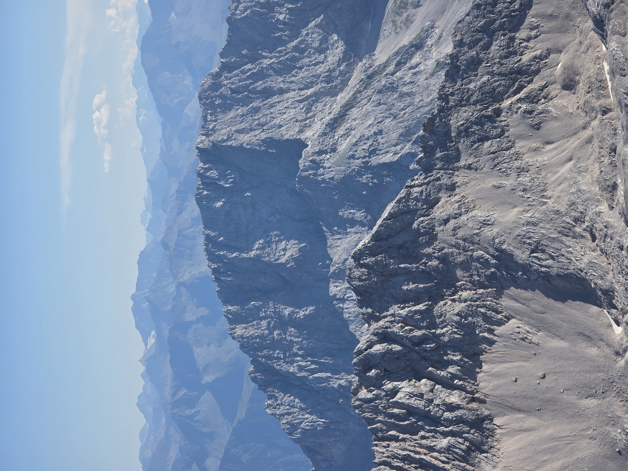 The image is an aerial view of the Zugspitze mountain range near Grainau, Germany. The foreground features jagged, rocky mountain peaks with slopes covered in scree and sparse vegetation. In the midground, a larger, imposing mountain dominates the scene, its sheer rock face deeply carved by erosion. The background reveals a series of receding mountain ranges fading into a hazy blue, indicating their distance. The sky above is a clear, pale blue with a few wispy clouds. There are no people or man-made objects visible in the image. The overall impression is one of rugged, natural grandeur and vastness.