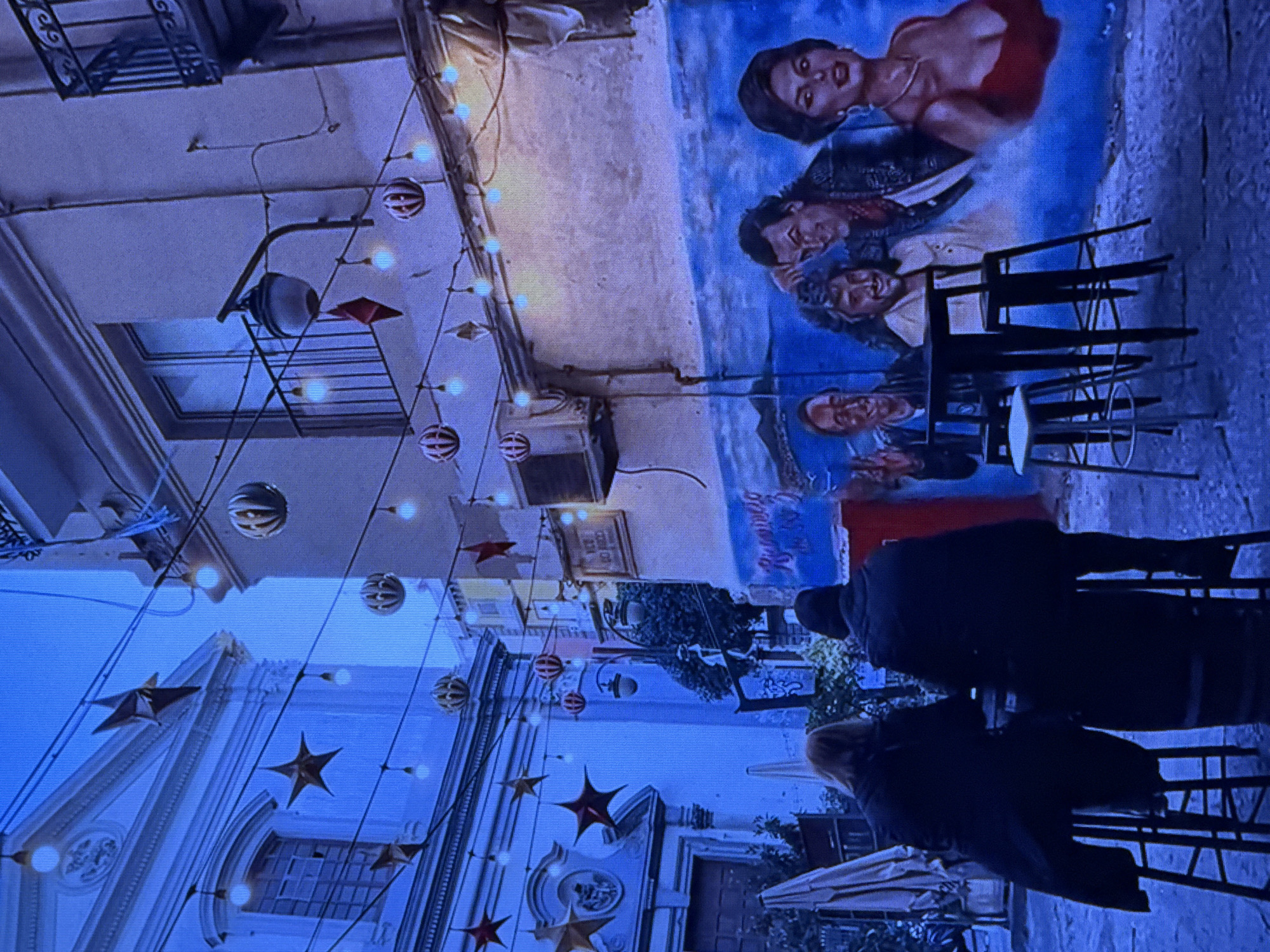 The image depicts an outdoor seating area with two individuals seated at a tall table, facing away from the camera. They are positioned in front of a mural painted on a building. The mural features several portraits of people. The scene is adorned with string lights and decorative stars. To the left, a portion of a building with architectural details and a window is visible. The overall lighting suggests it is evening or twilight, with a blue hue casting over the scene. There is no indication that this location is in Dubai, United Arab Emirates.