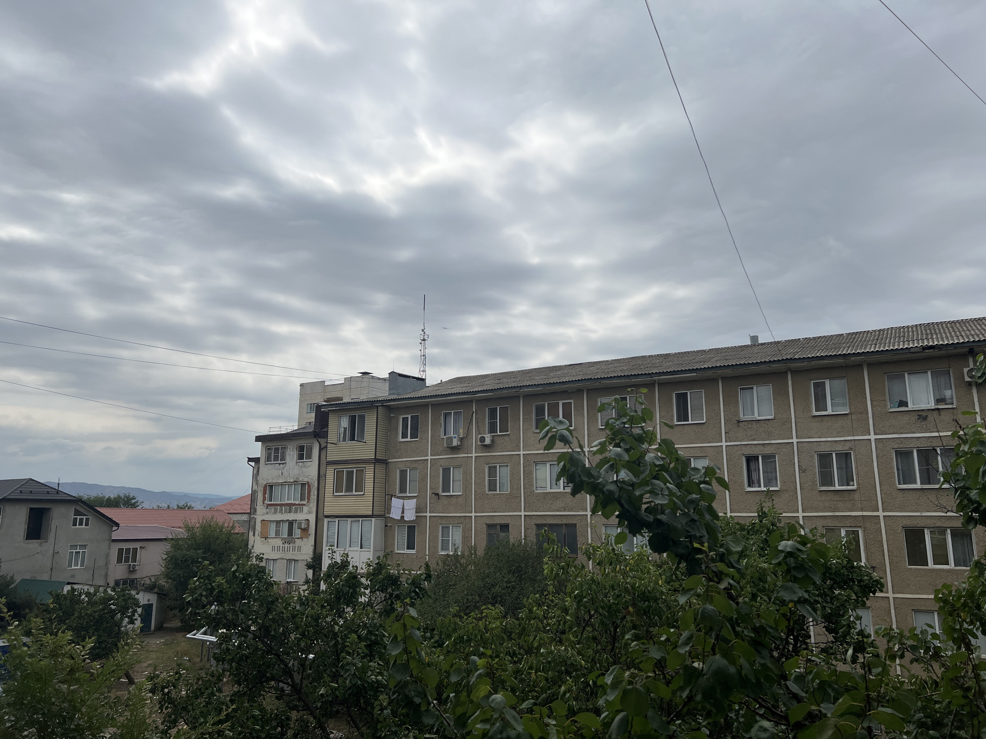 The image depicts a cloudy, overcast sky over a residential area in the city of Kizilyurt, Russia. The foreground is dominated by dense green foliage from trees and bushes. Behind the vegetation, several apartment buildings are visible. The central apartment building is a multi-story structure with a grey facade and numerous windows. To its left, a section of a building with a balcony and lighter colored panels is attached. Further to the left, other residential buildings with different architectural styles and roof types are present. In the background, distant hills or mountains are faintly visible through the haze. Power lines crisscross the sky. A telecommunications tower with antennas can be seen in the distance. The overall impression is a typical urban residential scene under a grey sky.