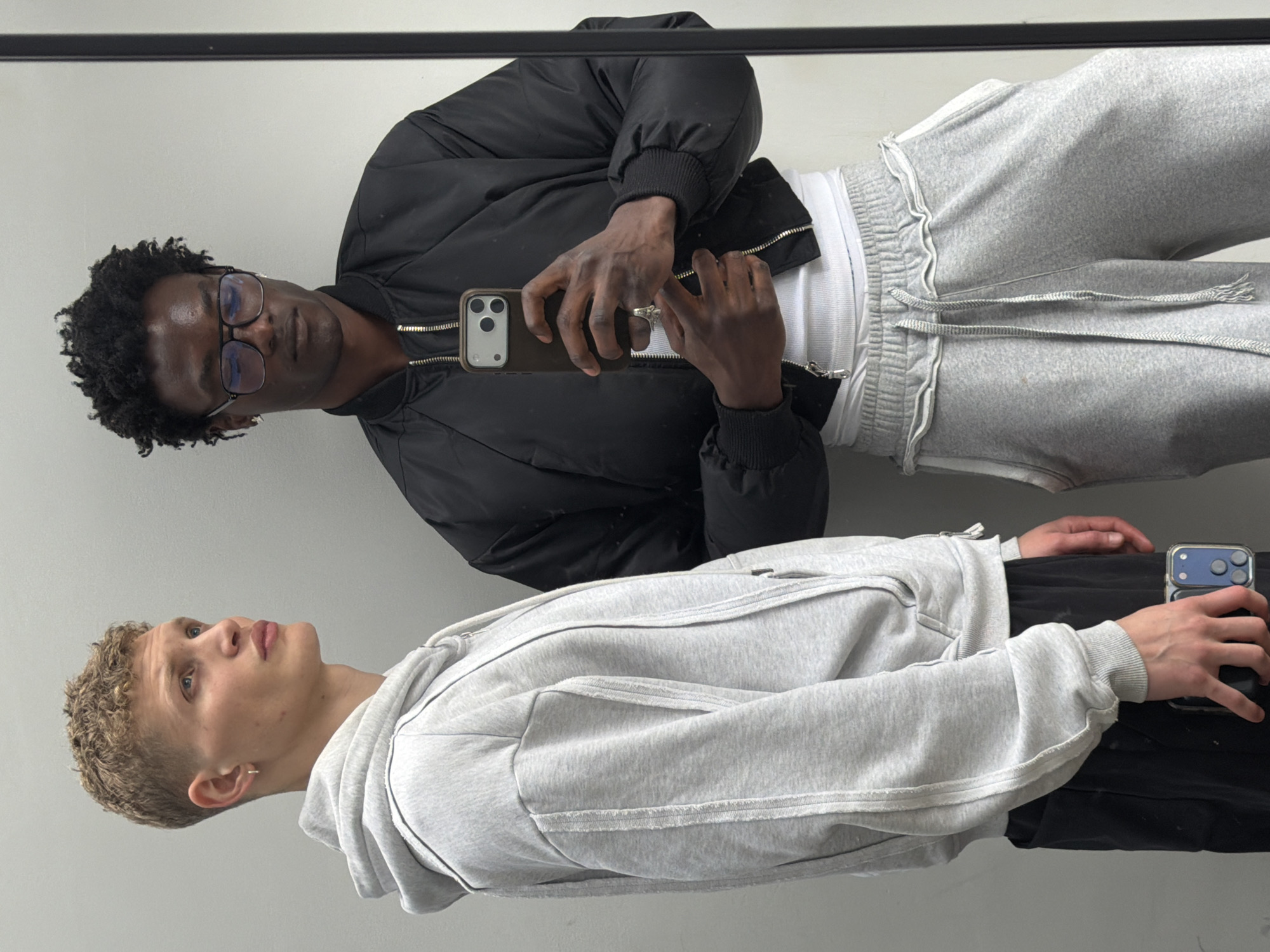 This media file is a mirror selfie featuring two young men, likely taken indoors against a plain, light-colored wall. A dark, horizontal bar, possibly a mirror frame, runs across the top of the image.
On the left, a man with dark skin, short, curly black hair, and glasses is seen. He is wearing a black bomber jacket over a white ribbed tank top or t-shirt, and light grey sweatpants with a visible drawstring. He holds a dark-cased smartphone with a light grey camera module in his right hand, seemingly taking the photo, and wears a ring on his right middle finger. His expression is neutral as he looks towards the camera.
On the right, another man with lighter skin, short, curly blonde hair, and an earring in his left ear is visible. He is dressed in a light grey zip-up hoodie featuring distinct stitching details and black pants or shorts. He holds a blue-cased smartphone with a black camera module in his hands. His gaze is directed slightly upwards and to his left, away from the camera.
Both subjects are reflected in the mirror, indicating an indoor setting. There are no specific location cues visible in the image itself to confirm the London context. No visible text is present.