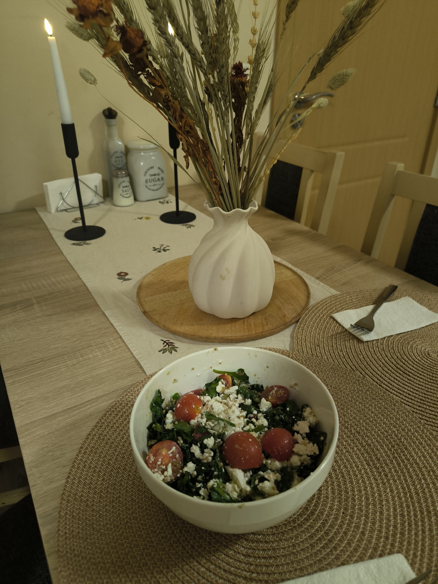 An indoor, eye-level shot captures a dining table set for a meal in an unknown city. The table, constructed of light-colored wood, is centered with a light beige fabric table runner adorned with small, stylized floral and leaf patterns.
In the foreground, a white ceramic bowl filled with a salad rests on a round, woven placemat of light brown material. The salad consists of dark green leafy vegetables, multiple whole and halved red cherry tomatoes, and crumbled white cheese. Visible behind the bowl is another identical woven placemat, featuring a small metal fork positioned on a folded white paper napkin.
Centrally placed on the table runner, on a circular wooden trivet, is a white, fluted ceramic vase holding a dried floral arrangement composed of various grasses, wheat stalks, and dried brown-orange floral elements.
To the left of the vase, on the table runner, a black metal candlestick holds a lit white taper candle, casting a warm glow. Behind it are a silver wire napkin holder with white paper napkins and a small white shaker labeled "SALT". To the right of the vase, another black metal candlestick stands, accompanied by a larger white ceramic jar labeled "SUGAR" with a dark lid, and a white bottle with a stopper, possibly an oil dispenser, labeled "OIL".
The background reveals a plain, light-colored wall to the left. To the right, light-colored wooden dining chairs with dark seatbacks are partially visible, suggesting a dining area. A light brown wooden door with a metallic handle is visible in the upper right background. No people are present in the scene.