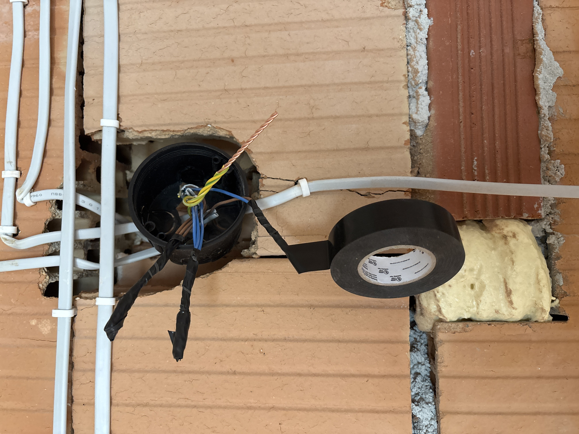 A close-up shows an exposed electrical junction box installed in a brick wall. The junction box is black and round, with several electrical wires extending from it. Some wires are capped with black electrical tape. A roll of black electrical tape is positioned to the right of the junction box, partially obscuring a section of the wall. White plastic conduits run along the wall to the left of the box. Spray foam insulation is visible in gaps between bricks. The location is Burgas, Bulgaria.