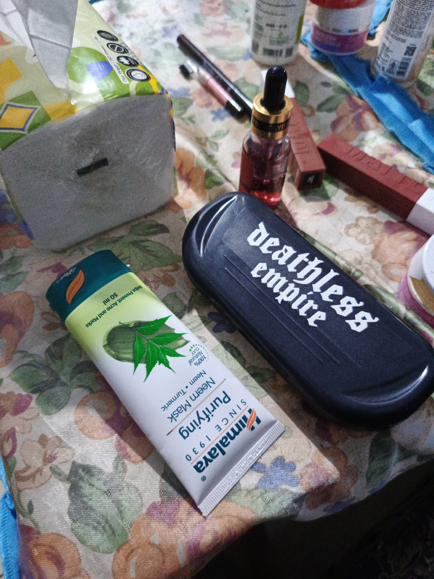 A flat lay image shows a collection of beauty and personal care products arranged on a floral patterned tablecloth. In the upper left is a partially visible package of tissues. To its right, a black pen with a pink accent lies horizontally. Below the pen, a small glass bottle with a dropper cap contains a red liquid, and next to it are two rectangular tubes, one red and one a lighter shade.
Dominating the center right is a black, oval-shaped case with "deathless empire" printed in white gothic-style lettering. In the lower center, a white tube of Himalaya Purifying Neem Mask is positioned diagonally. The tube displays an illustration of neem leaves and mentions "100% Natural Clay," "Neem," and "Turmeric." The text "Helps Prevent Acne and Marks 50 ml" is also visible. The background is a busy floral print tablecloth with colors of pink, orange, green, and blue. The lighting suggests an indoor setting, possibly a personal vanity or table. The image is taken from a top-down perspective. There are no people or animals present. The location of this scene within the city of Sumbersari, Indonesia is not identifiable from the visual information provided.