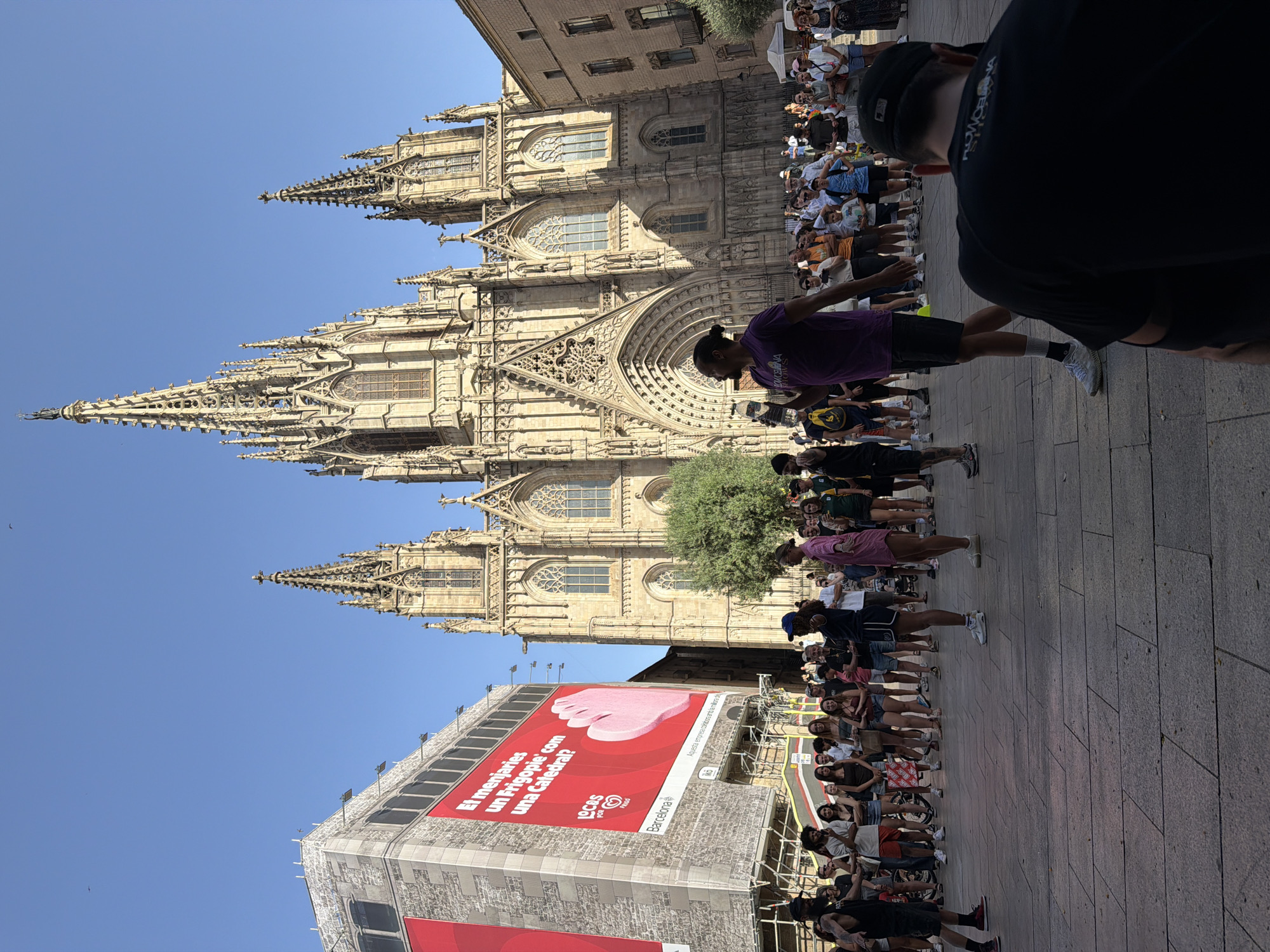 The image depicts an outdoor scene in front of the Barcelona Cathedral in Spain. A large crowd of people is gathered in a paved plaza. In the foreground, a person in a purple shirt and black shorts is facing away from the camera, holding a water bottle. To their left, a person wearing a black t-shirt and shorts is also facing away. In the middle ground, several people are standing and observing. A prominent building with a large red advertisement featuring a hand and text in Catalan is visible on the left. The Barcelona Cathedral, with its gothic architecture and spires, dominates the background. The sky is clear and blue. The overall scene suggests a public gathering or event.
