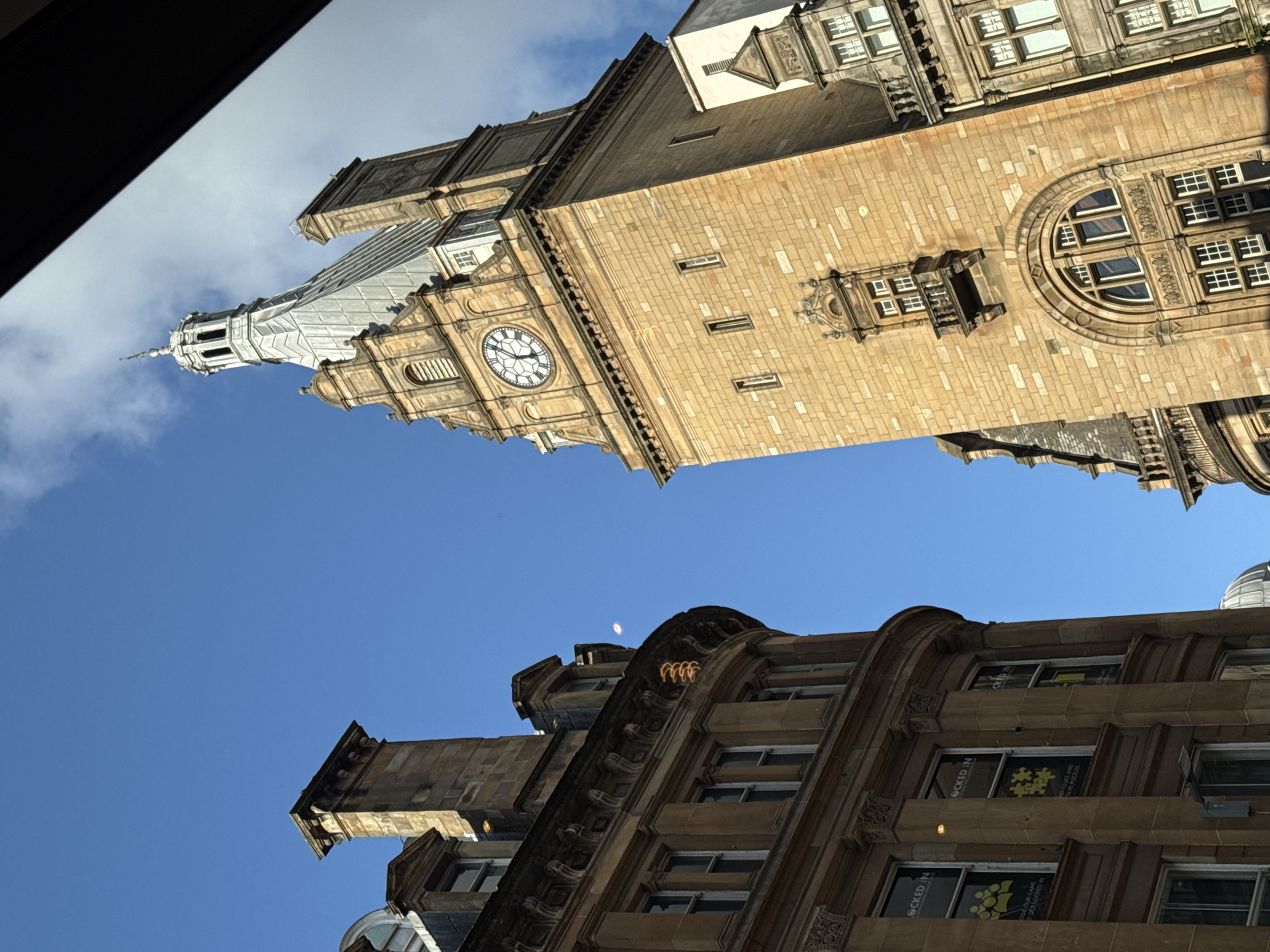 This is a low-angle, slightly tilted photograph of the iconic Glasgow City Chambers clock tower, viewed from the street. The tower is constructed of warm, sandstone blocks and features a large, white clock face with black Roman numerals. Above the clock, the tower narrows and culminates in a silver, spire-like roof with a weather vane. The sky behind it is a clear, bright blue, with a few wisps of white clouds.
To the left of the clock tower, the curved facade of another building is visible. This building is also made of sandstone and has multiple windows with white frames. Some of the windows have posters displayed inside, which appear to be for an escape room called "Locked In." The architectural style of this building is ornate, with decorative moldings and cornices.
The overall scene depicts a sunlit urban environment with impressive historical architecture. The vantage point suggests the photograph was taken from street level, looking upwards at the grand buildings. There are no people or vehicles visible in the frame. The lighting indicates it is daytime, likely late morning or early afternoon given the angle of the sun.