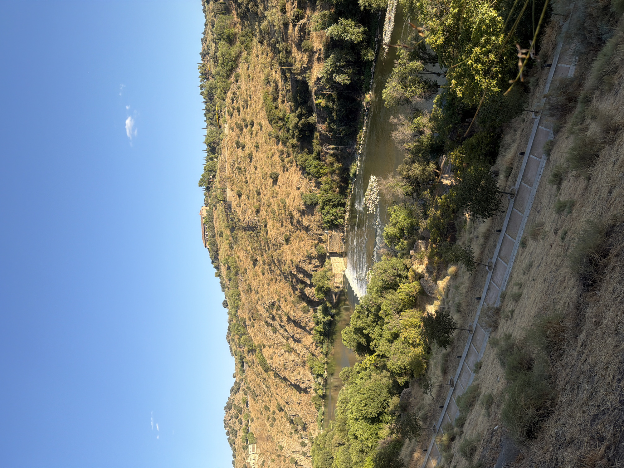 This is a wide, landscape photograph of the Tagus River in Toledo, Spain, taken on a clear, sunny day. The river is bordered by lush green trees on both banks. A small waterfall is visible in the river, and a stone structure, possibly part of an old dam or bridge, is situated behind it. The hillsides surrounding the river are covered in dry, golden grass and dotted with scattered trees. In the distance, on top of one of the hills, a large building with a red-tiled roof is visible. A paved pathway with several young trees lining it runs along the grassy slope in the foreground, leading the viewer's eye towards the river. The sky is a vibrant blue with a few small, wispy clouds.