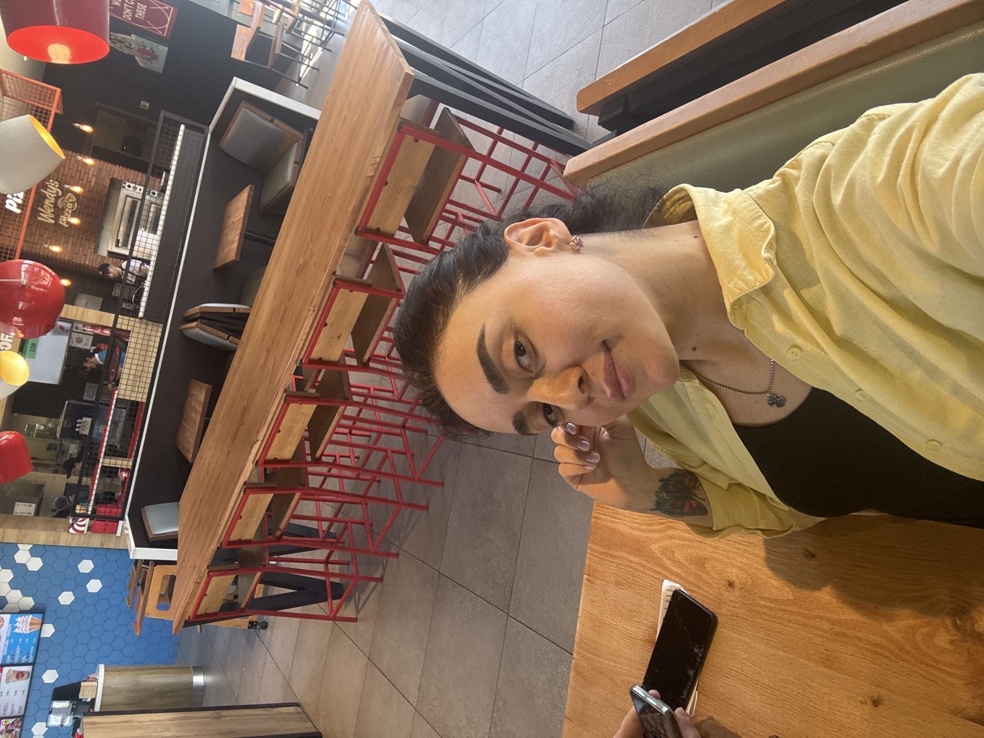 The image shows a woman taking a selfie at what appears to be Wendy's Pizza restaurant in Tbilisi, Georgia. The restaurant interior is visible, with long wooden tables and red metal frames. The woman is wearing a yellow button-down shirt over a black top. A smartphone with a cracked screen sits on the table next to her. In the background, there are tables and chairs, along with signs for Wendy's Pizza. The blue and white hexagonal floor tiles are visible near the entrance.