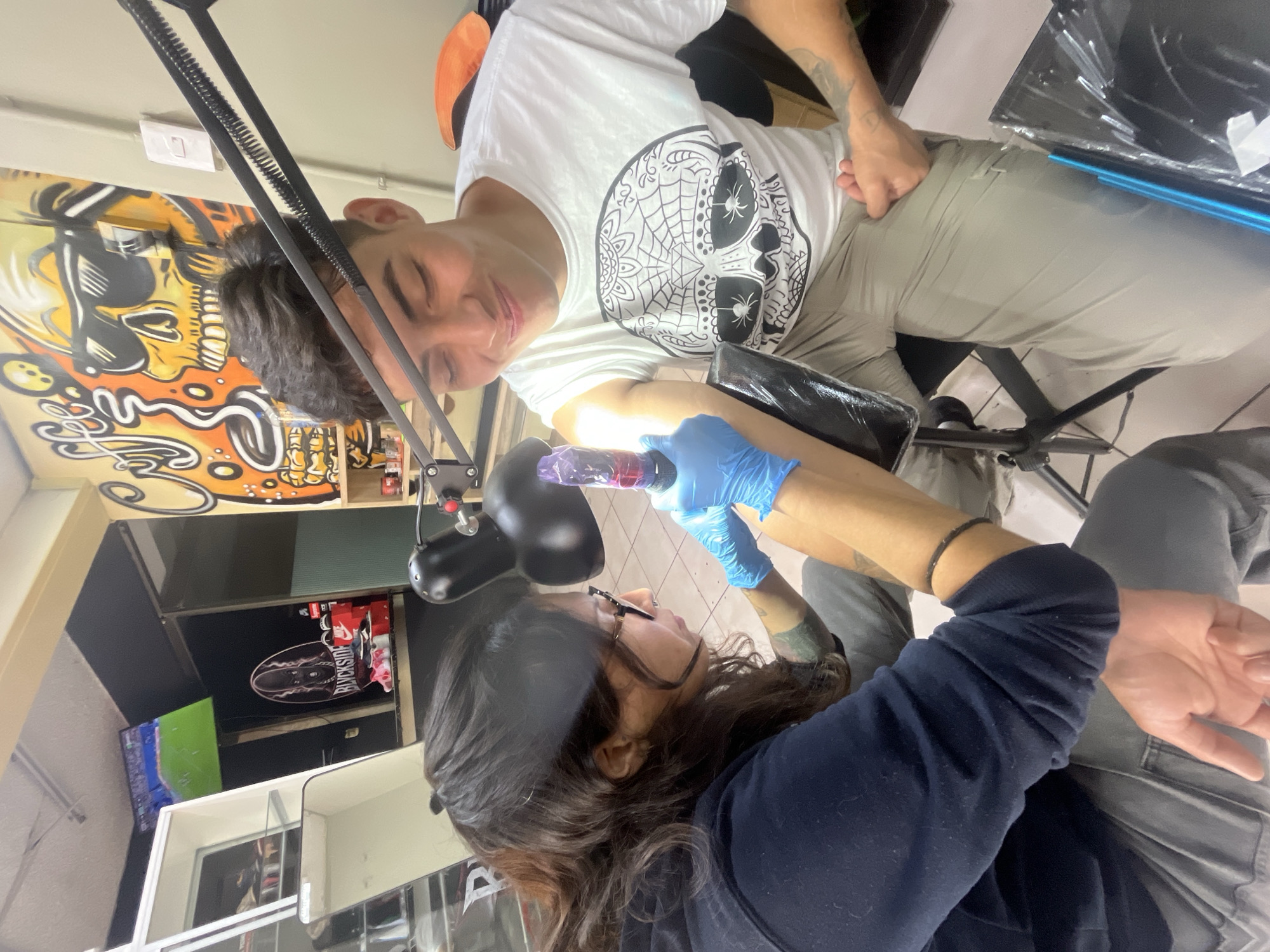 In a tattoo parlor located in the city of Iquique, Chile, a tattoo artist with long, dark hair and glasses, wearing a dark blue hoodie and blue gloves, is in the process of tattooing a young man. The artist holds a tattoo gun to the man's upper arm. The man, who is wearing a white t-shirt with a black sugar skull design and light-colored pants, is seated and looking down with a slight smile. The environment appears to be a commercial space, indicated by a display shelf with shoes and merchandise in the background, as well as a television screen showing a sporting event. A large, colorful mural of a skull is visible on the wall behind the man. The scene is lit by a bright lamp positioned over the artist's work area.