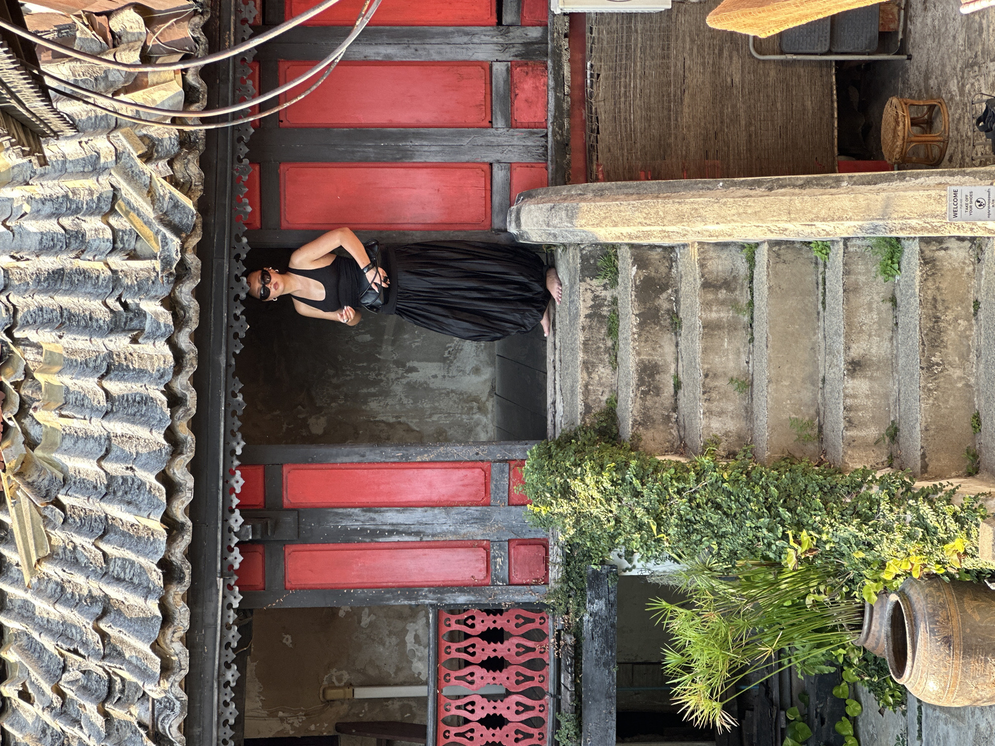 A woman in a black dress stands between an interior hallway with red and black lacquered wood walls and a flight of concrete steps leading upward. She wears sunglasses and holds black heeled sandals. The location is near Wat Pho in Bangkok, Thailand, identified by the traditional Thai architectural style of the building. The hallway features a tile roof overhang and ornate woodwork. The steps are lined with lush green vegetation growing out of a large earthenware pot. A sign reading "WELCOME - TAKE OFF YOUR SHOES" is visible near the top of the steps.