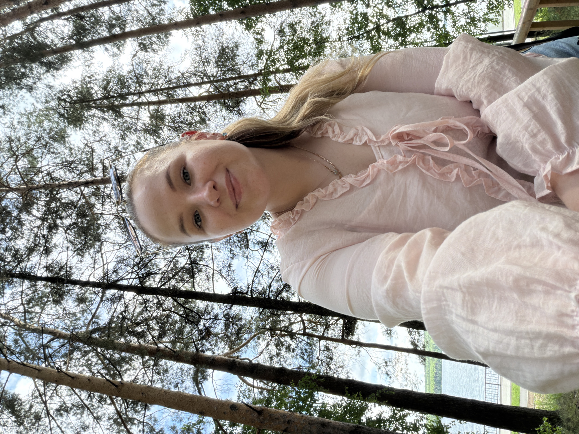 A young woman with blonde hair pulled back in a ponytail and wearing a light pink ruffled blouse is taking a selfie outdoors. She is framed by tall pine trees, with a body of water visible in the background. She is smiling gently at the camera, and her necklace is visible. The setting appears to be a park or recreational area, possibly near a lake or river. The scene is set during daytime, under a cloudy sky.
