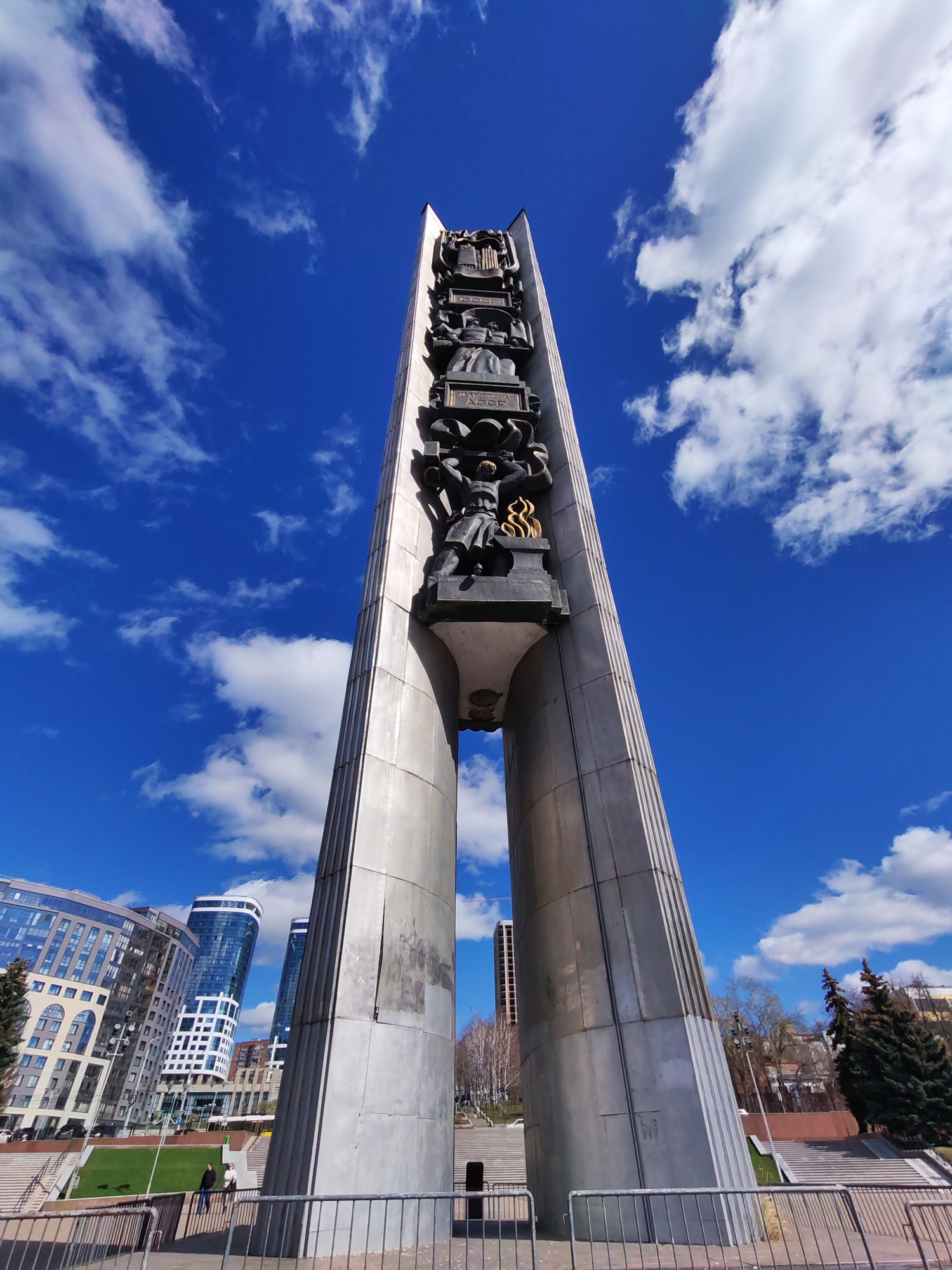 A low-angle, wide-angle shot depicts the Monument of Eternal Fire in Izhevsk, Russia, against a vibrant blue sky dotted with puffy white clouds. The monument, constructed of a gray, metal-like material, rises high, with a vertical, sculpted centerpiece showing figures.
In the background are modern city buildings, and trees line the landscape. A gray metal fence is visible in the foreground.