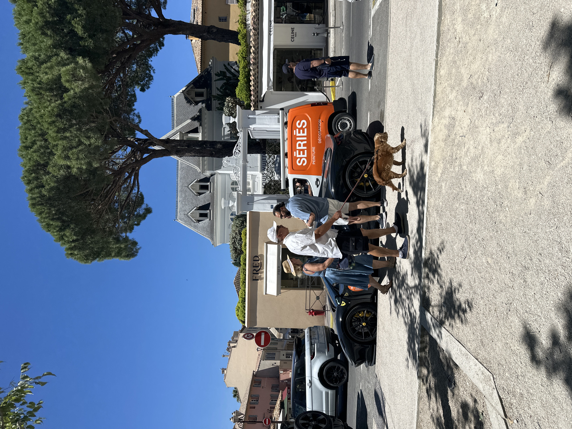 The image depicts a street scene in Saint-Tropez, France, on a bright, sunny day. In the foreground, a black sports car is parked on the side of the road. A man and a woman are standing next to the car, and a small, golden-brown dog on a leash is walking in front of them. The man is wearing a white cap and a white short-sleeved shirt, with black shorts. The woman is wearing a blue dress and a straw hat.
Behind the sports car, a white Range Rover is parked. Further down the street, an orange van with "SÉRIES PEINTURE DÉCORATION" written on its side is visible. Several shops with awnings line the street, including "FRED" and "CELINE." People are walking on the sidewalk, and more buildings with characteristic tiled roofs are visible in the background. Trees with lush green foliage provide shade. The sky is clear and blue.