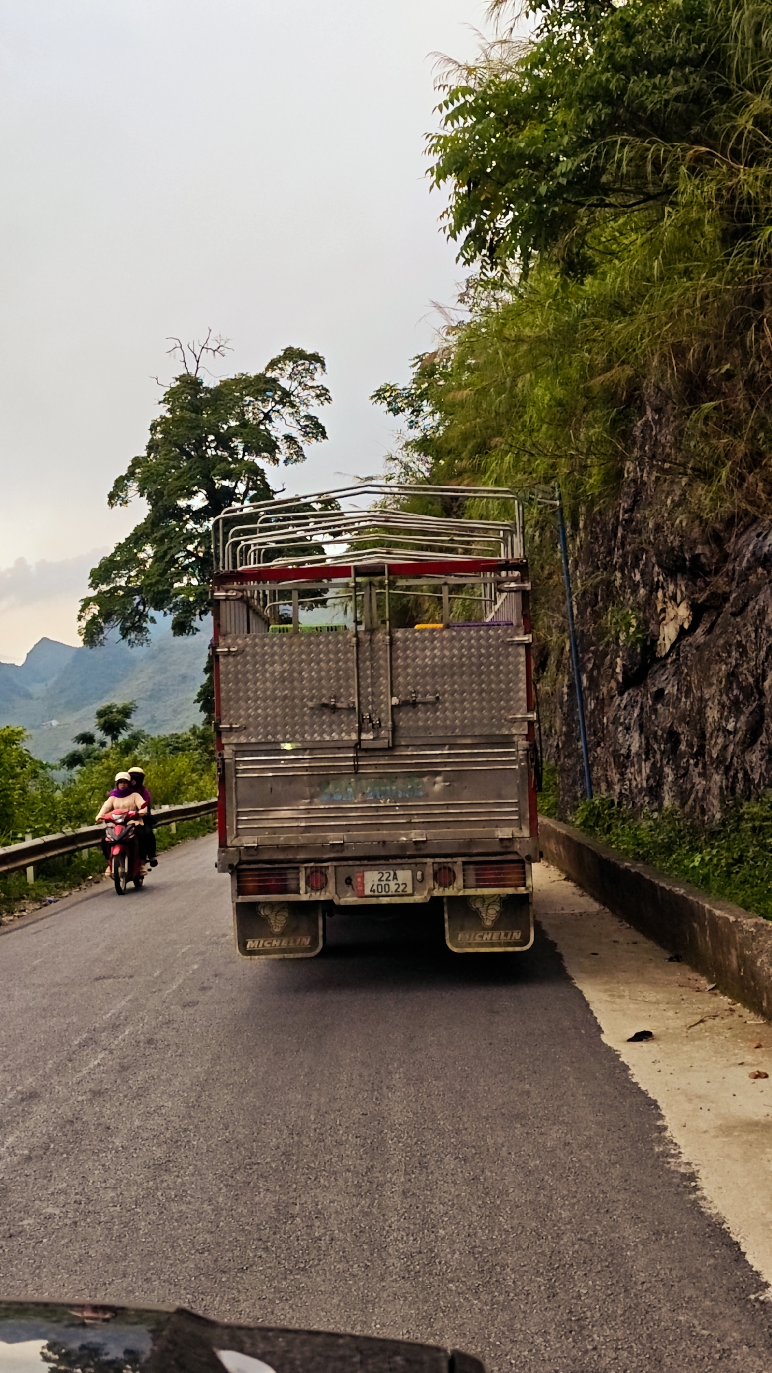 The image depicts a road scene in Cán Tỷ Commune, Vietnam. A medium-sized truck with a cargo bed dominates the center of the frame, driving on a narrow road. Behind the truck, a motorcycle with two riders is approaching. To the right of the truck is a rocky embankment with green vegetation. The road itself appears well-maintained. In the background, a mountainous landscape is visible under a cloudy sky. A distinctive tree stands out against the backdrop.