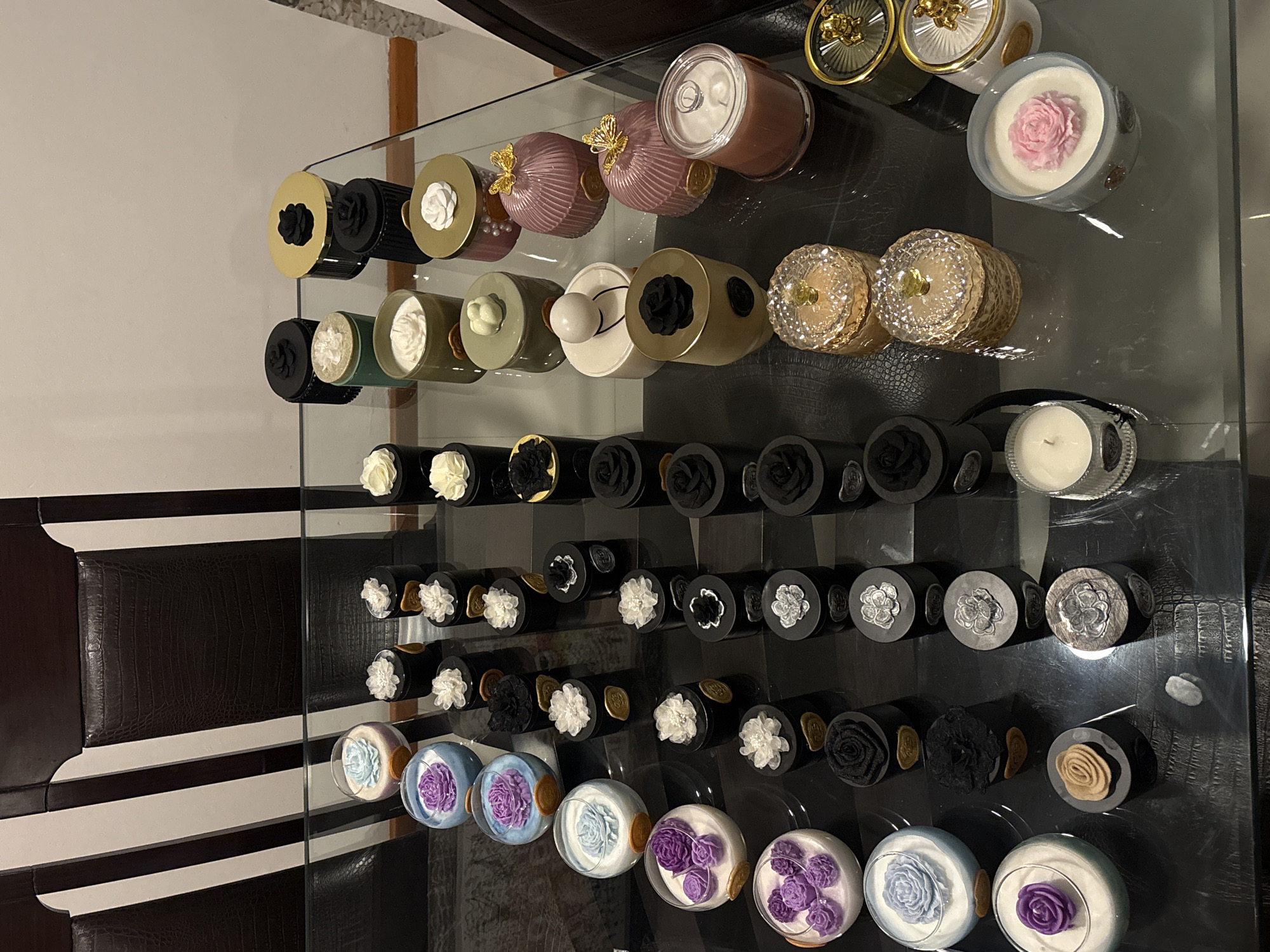 The image shows a collection of decorative candles arranged on a glass table. The candles are in various containers, including cylindrical jars and round glass bowls, with lids or decorative tops. The designs on the candles include floral motifs, such as roses and other stylized flowers, in a range of colors including black, white, gold, pink, purple, and blue. The background features a wall and a piece of furniture with a dark, patterned design.
There is no information to indicate the specific location of the city of San Bernardino Tlaxcalancingo, Mexico.