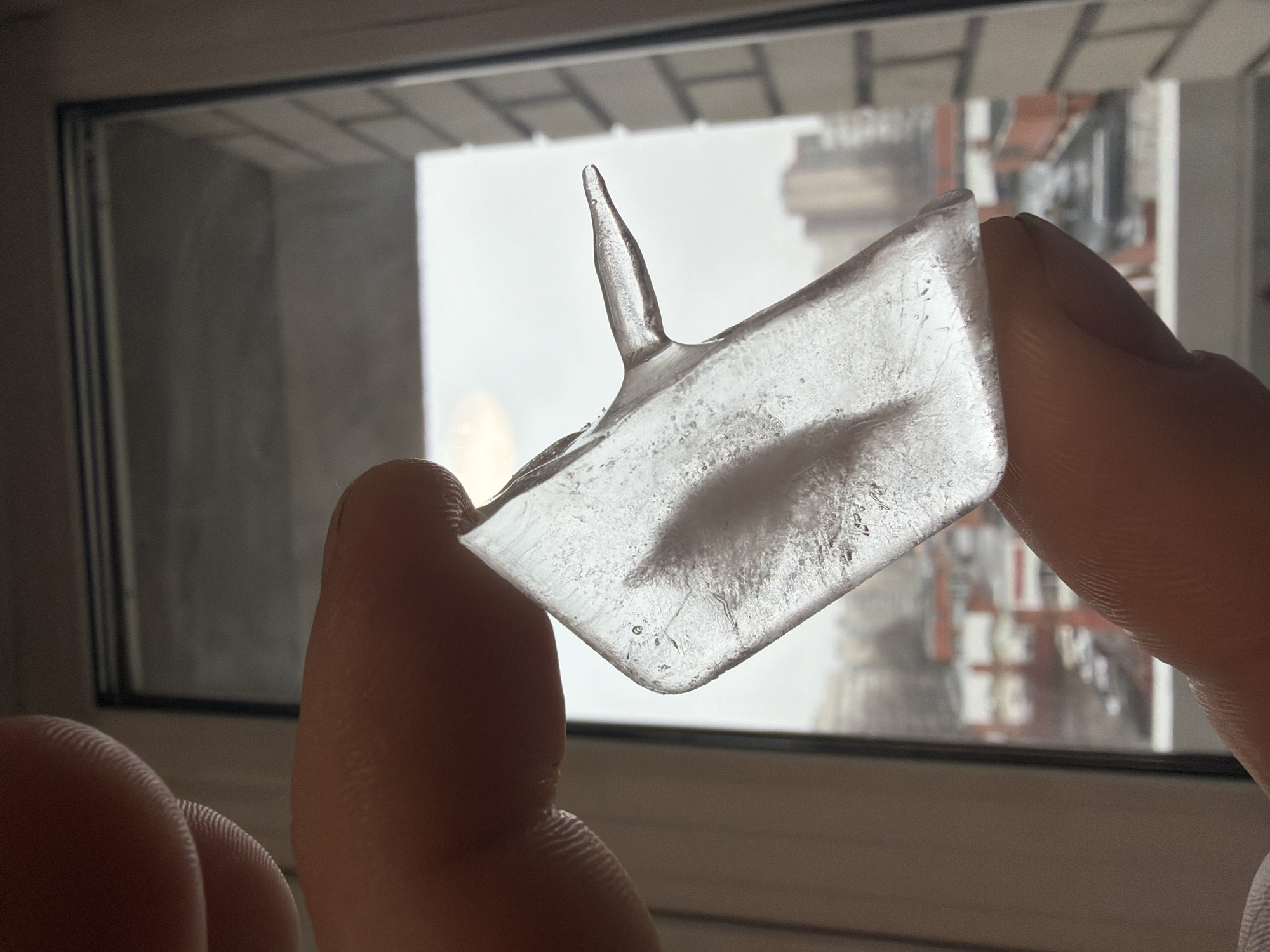 A person holds a translucent, roughly rectangular shard of ice with a thin spike extending upwards. The ice shard is held between the thumb and forefinger. The background is a window, offering a blurred view of a cityscape that includes buildings with red and white facades. Based on the architectural style visible through the window, the location is the city of Saint Petersburg, Russia.