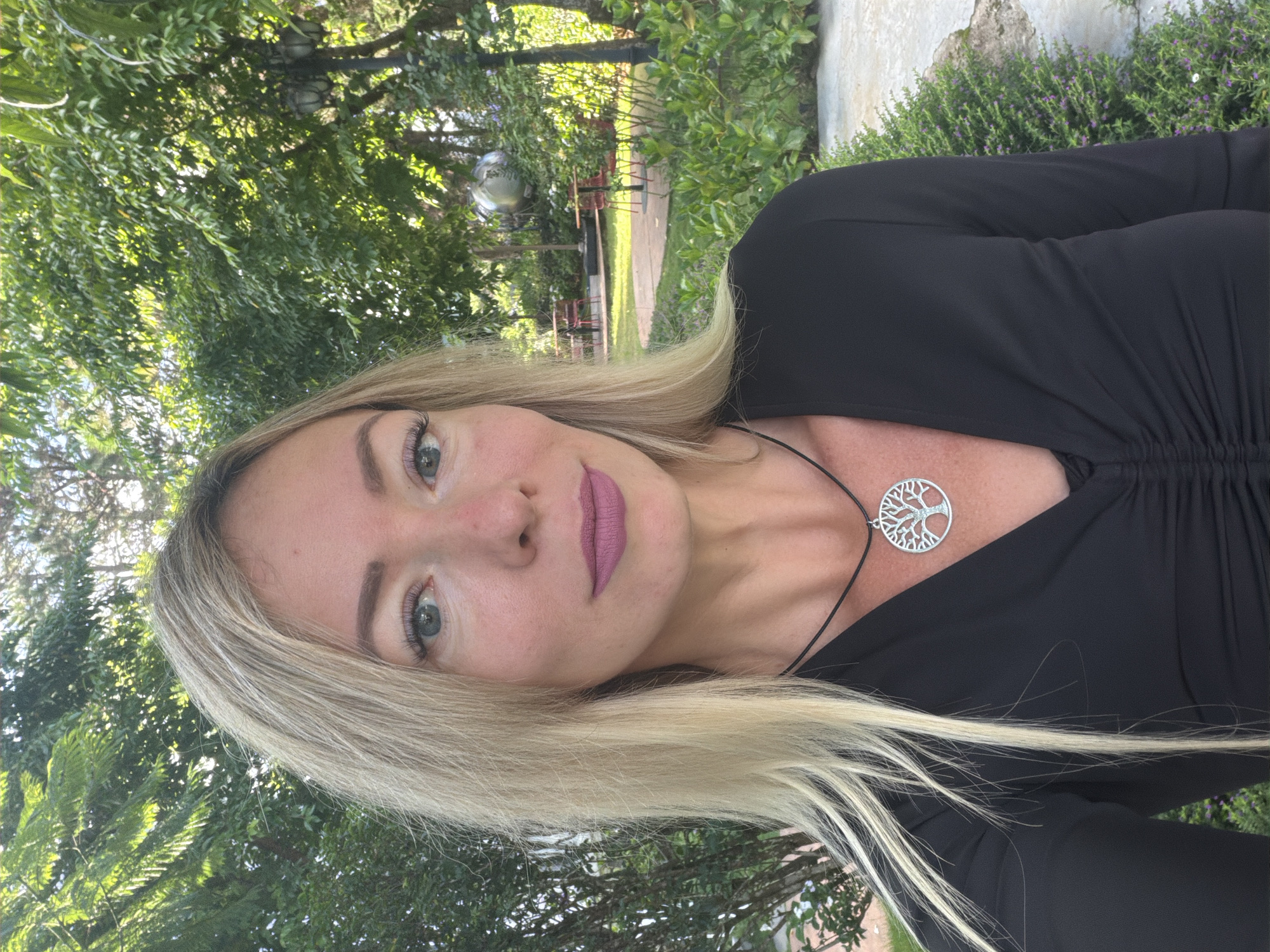 A woman with blonde hair and light blue eyes is pictured in an outdoor setting. She is wearing a black, long-sleeved shirt with ruching across the bust and a silver pendant shaped like a tree of life on a black cord. Her makeup includes pink lipstick and mascara. The background suggests a garden or park with lush green trees and plants, some visible tables and chairs, and a spherical sculpture. The lighting indicates it is daytime. The location is identified as Xuân Trường - Đà Lạt, Vietnam.