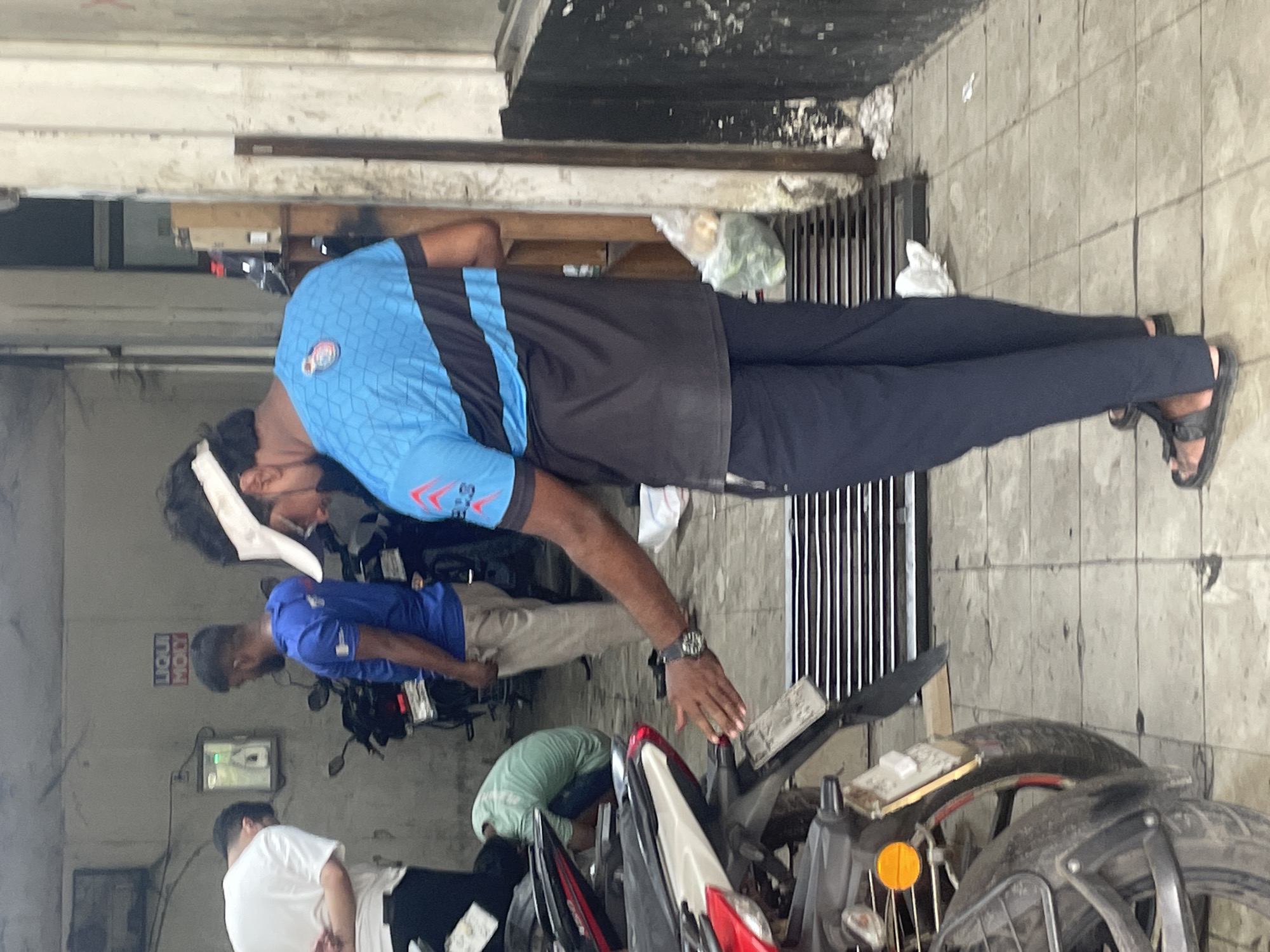The image shows a group of individuals in what appears to be a motorcycle repair shop in Dhaka, Bangladesh. One man wearing a blue and black shirt and dark pants is inspecting a motorcycle. Another man in a blue shirt is working on a motorcycle. A third man in a white shirt and black pants stands nearby. A fourth person, wearing a green shirt, is crouched down near the motorcycles. There are several motorcycles visible, some in various stages of repair. The shop appears to be located indoors.