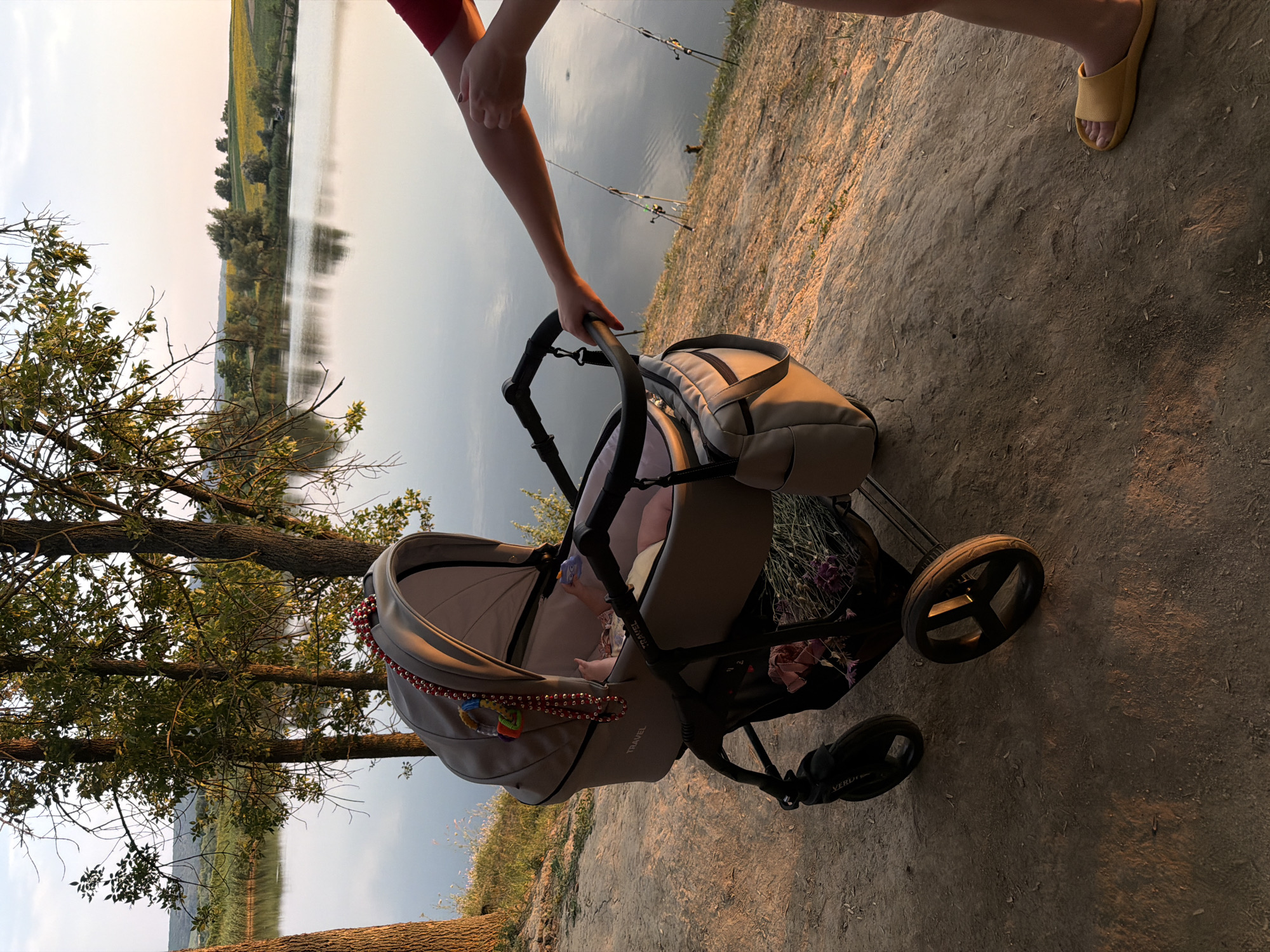 A baby is in a gray stroller on a dirt path next to a lake. The stroller has a red and white beaded garland hanging from the canopy, and a toy attached to it. The baby's feet are visible. A person's arm is holding the stroller handle. There are fishing rods in the background by the lake. Trees and fields are visible across the lake. The sun is setting, casting a warm glow on the scene. The location is Strășeni, Moldova.