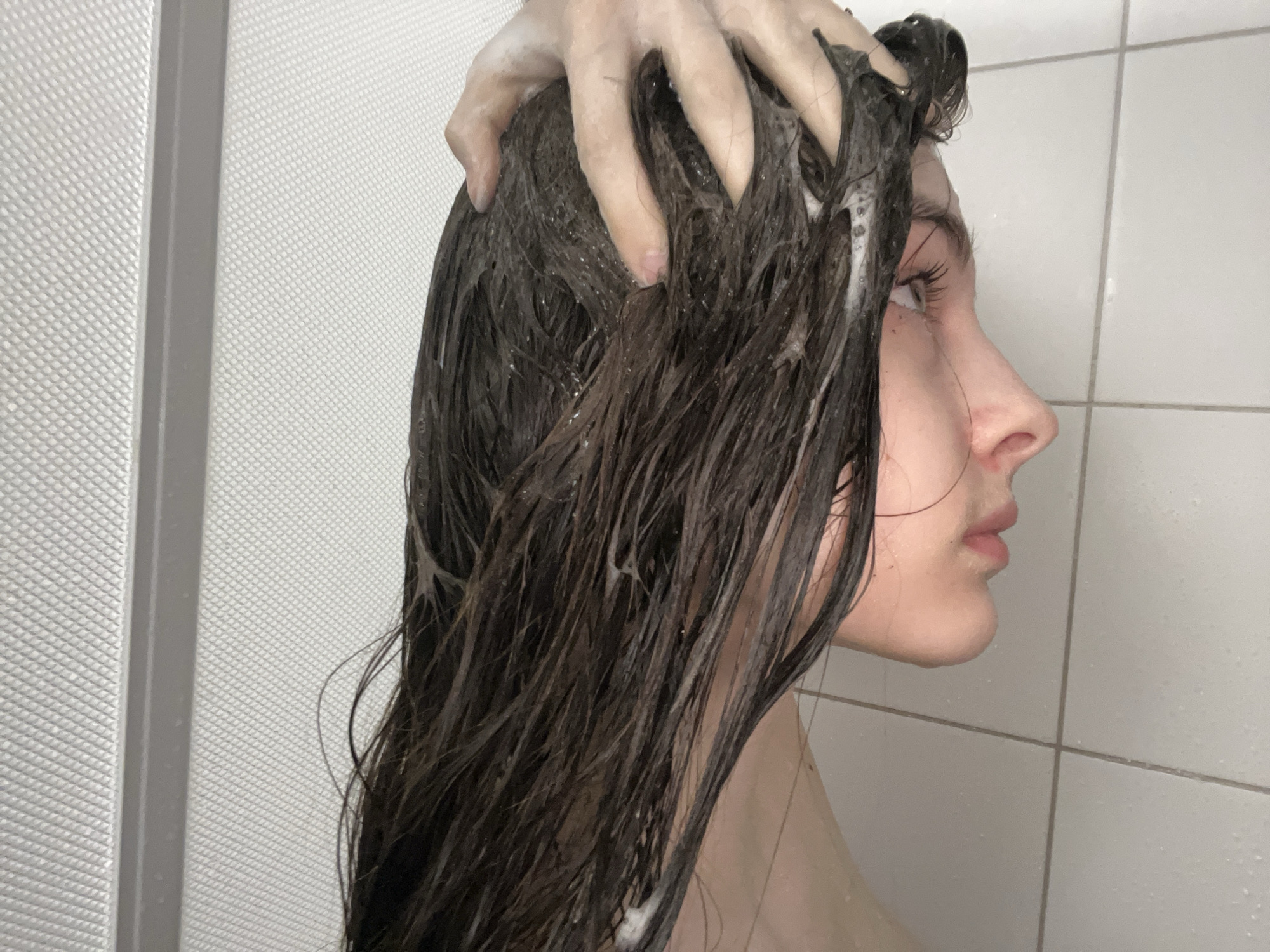 A person is in a shower washing their hair. The person's hair is long and dark, and is covered in soap or shampoo. The person is using their hands to scrub their scalp. The shower has white tiled walls. The location is not discernible as Saint Petersburg, Russia.