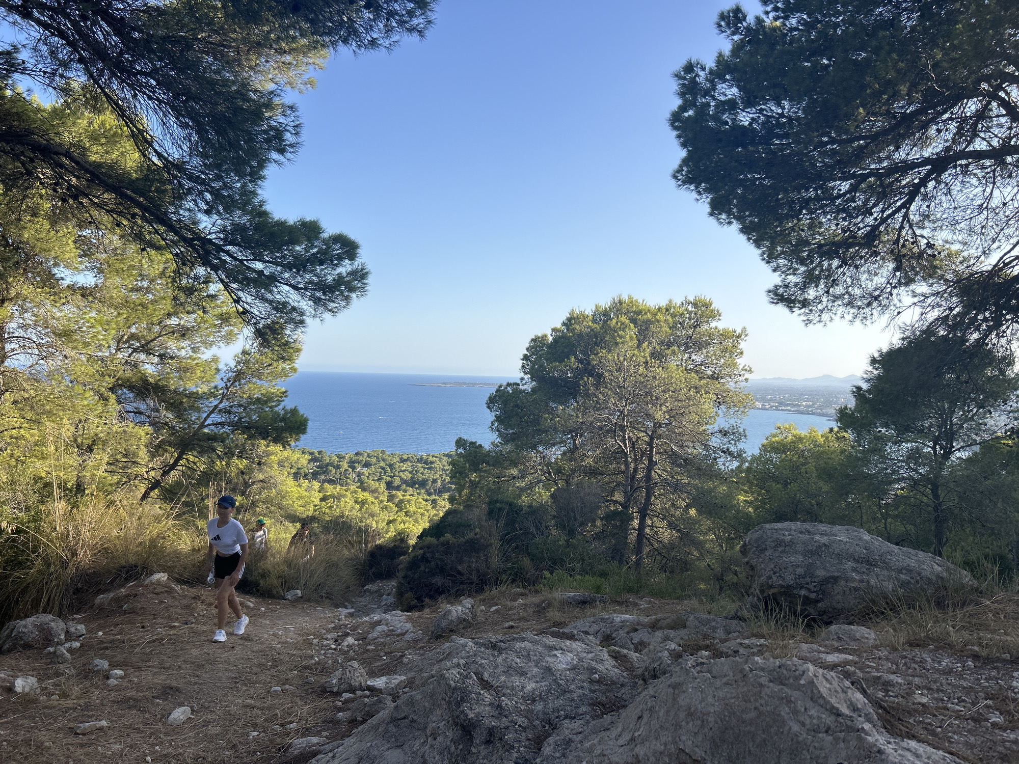A full shot captures a person walking down a rocky, dirt path in a wooded, hilly area overlooking a body of water and a coastal town. The person, seen from the side and slightly from the front, is wearing a white t-shirt, black shorts, white sneakers, and a blue baseball cap. They are carrying a water bottle in their right hand. Behind them, another person is visible, also walking down the path. The surrounding environment is characterized by dry grasses, scattered rocks, and pine trees. The sun is shining, casting shadows on the ground. In the background, a vast expanse of blue water is visible, meeting the horizon under a clear blue sky. Beyond the water, a town or city with buildings can be seen along the coast, with hills or mountains in the distance. The location is identified as Costa Des Pins, Spain.