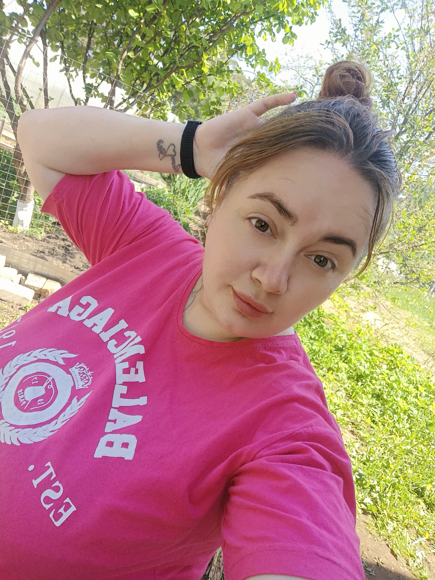 A woman with brown hair tied in a bun is posing for a selfie outdoors. She is wearing a pink t-shirt with the word "BYTECJAY" printed on it, along with some other lettering and a crest design. She has a tattoo and a black wristband on her left arm. The background includes green grass and trees, and a section of a wire fence and building materials. The location is Chaikovsky, Russia, judging by the text on the woman's shirt.