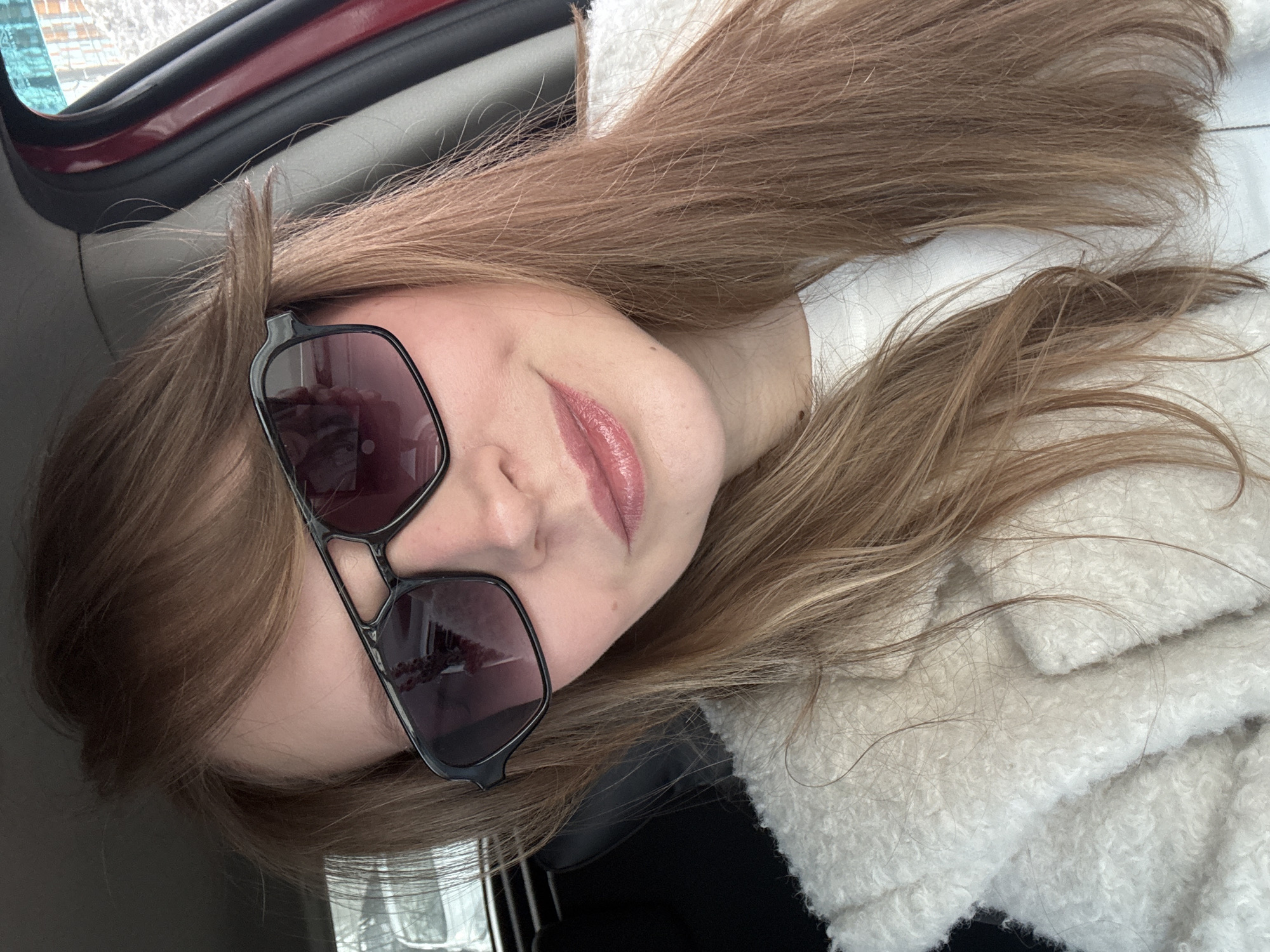 A young adult female is depicted in a close-up selfie taken from inside a vehicle in the city of Izhevsk, Russia. She has fair skin, long light-brown hair styled with bangs, and wears dark-tinted sunglasses with a black, angular, double-bridge frame. Her lips are slightly parted in a subtle smile, enhanced with a glossy pink lip product. She is dressed in a white garment underneath a cream-colored, textured, "teddy bear" style coat or jacket. The vehicle's interior features include a dark headliner and dark seat upholstery visible to the left of the subject. A red vehicle door frame and pillar are visible on the right side of the frame. Through the vehicle's windows, a blurred exterior environment is discernible, consistent with a winter scene featuring snow, faint outlines of buildings, and other vehicles. Reflections of the exterior environment are visible on the surfaces of the sunglasses lenses.