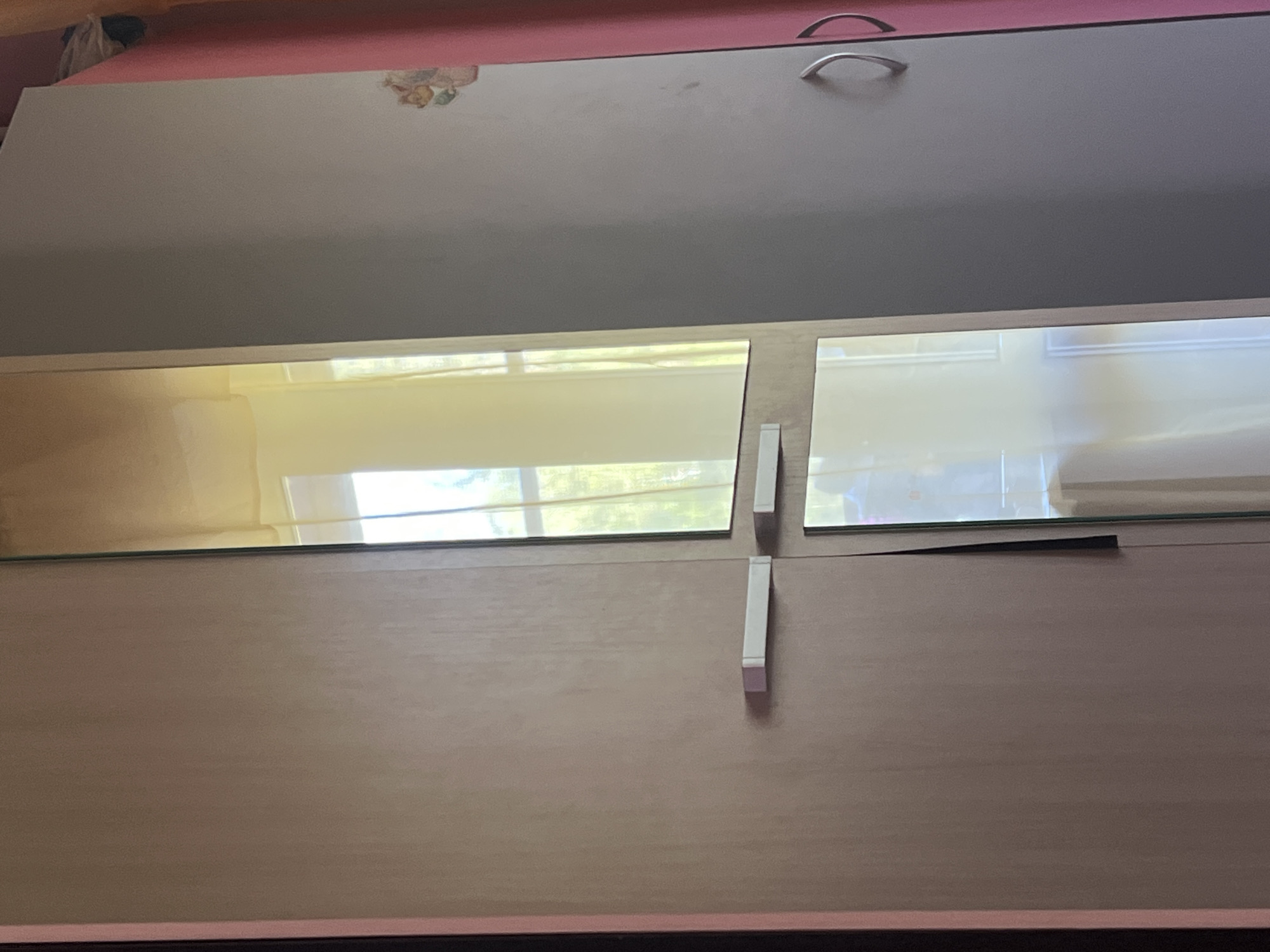 A low-angle, indoor shot depicts a section of a piece of furniture, likely a cabinet or dresser. The top portion is a muted gray with a silver, curved handle visible in the upper right. A small, blurred decorative element is present in the upper left of the gray section. Below the gray top, the furniture paneling has a light-colored wood grain pattern. Two rectangular glass panels are placed horizontally, meeting in the center. The left glass panel reflects a view of a window with a partial view of greenery outside. White supports are positioned at the junction of the glass panels and below the right panel. The lower edge of the furniture is lined with a dark trim. The color palette is dominated by shades of gray, brown, and white. No people are visible. No information about Noviy, Russia can be extracted from the image.