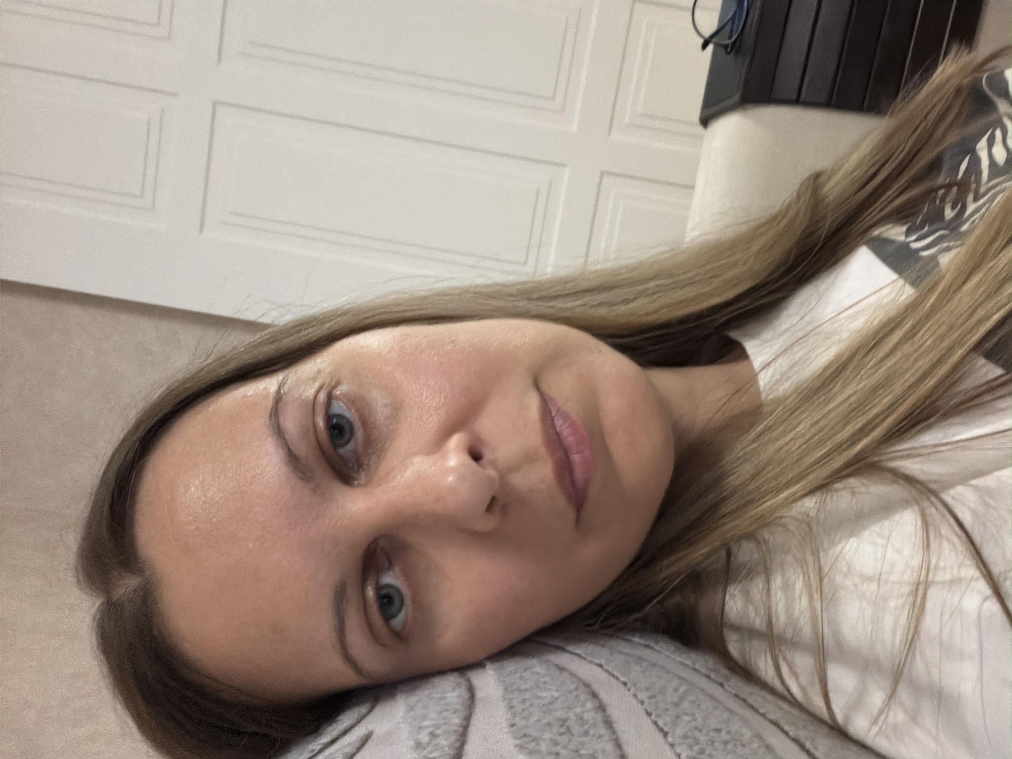 A woman is lying down and looking at the camera. She has shoulder-length blonde hair and blue eyes. She is wearing a white t-shirt. She is resting her head on a textured pillow. In the background, there is a white door and a dark chest of drawers with a pair of glasses on top. The lighting is soft and warm, suggesting an indoor environment during the day. The image appears to be a selfie, taken from a close-up perspective.