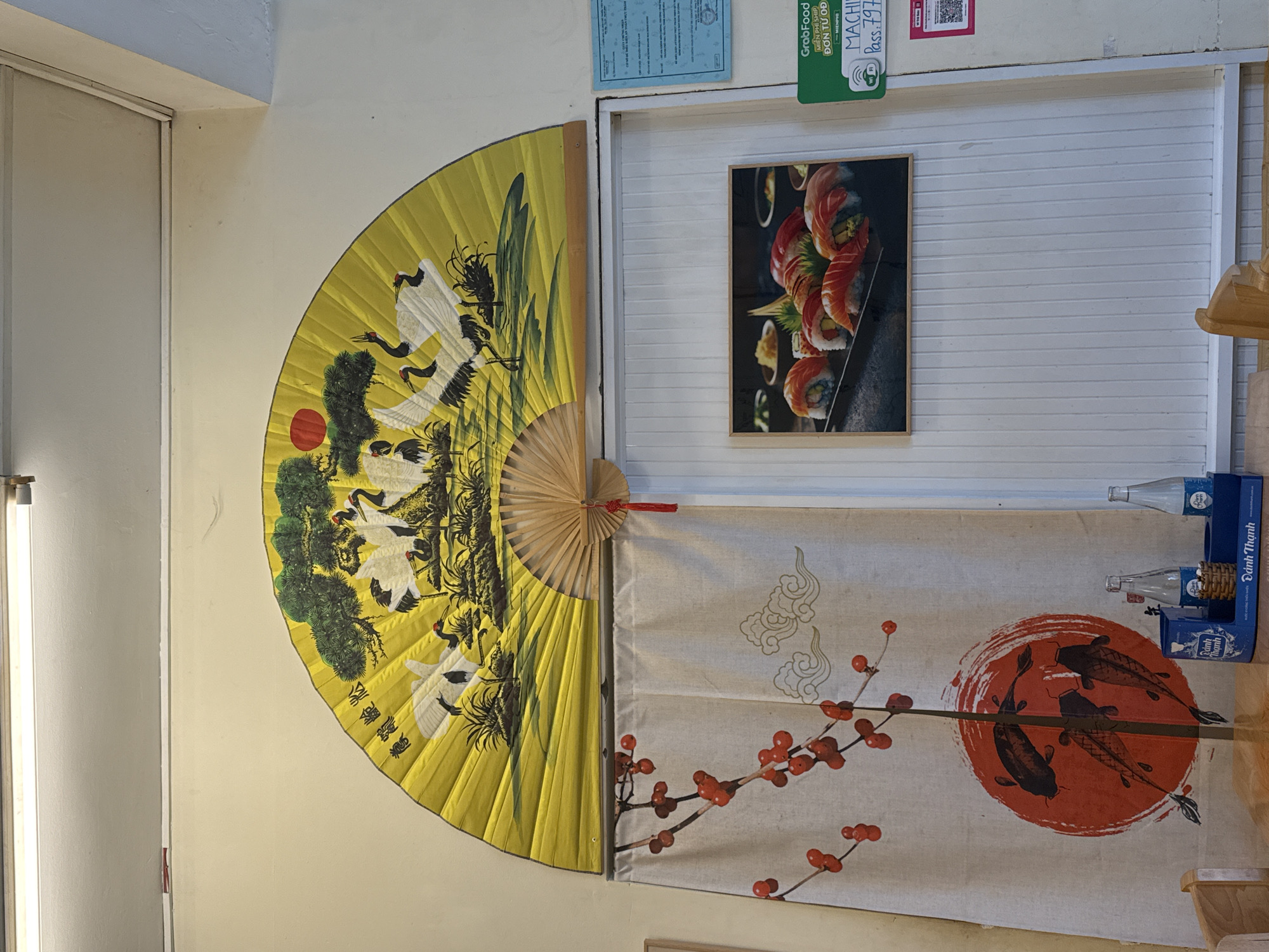 This is a forensic, matter-of-fact description of a restaurant interior in Nha Trang, Vietnam. The scene is illuminated by a fluorescent light fixture overhead. Dominating the upper portion of the wall is a large, open yellow fan decorated with a painting of several white cranes, pine trees, and a red sun. Below the fan, a cream-colored curtain hangs, featuring a design of red berries on branches and a circular emblem with three dark koi fish. To the right of the curtain is a framed photograph of sushi rolls. Further to the right, on the wall, are two blue official-looking documents and a green GrabFood promotional sign. A white, ribbed wall panel is visible behind the framed photo and the GrabFood sign. On a wooden table in the foreground, two clear glass bottles and a blue container holding more bottles are visible. The table is made of wood, and a portion of a wooden chair is also visible. The environment suggests a restaurant with an East Asian theme.