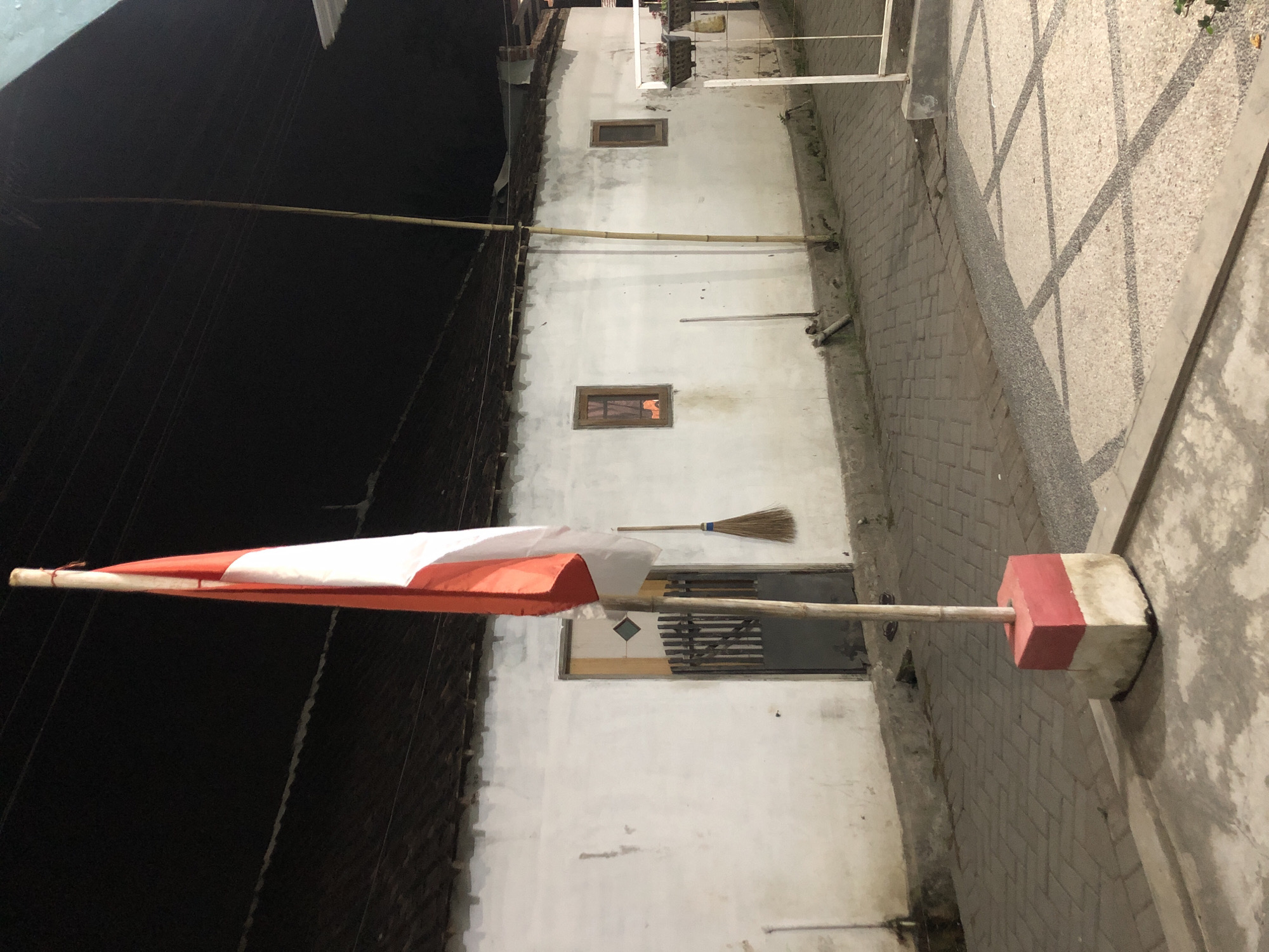 The image depicts an Indonesian flag flying from a bamboo pole in what appears to be a residential area at night. The flag, with its red and white horizontal stripes, is partially illuminated. The pole is secured in a concrete base painted red and white. Behind the flag, a white stucco wall with a dark-tiled roof suggests a building, possibly a house. A wooden door with a metal grate is visible on the wall, along with a broom leaning against it. Further down the wall, two rectangular windows are present. The ground in front of the wall is paved with interlocking bricks, and a patterned concrete area is visible in the foreground. A bamboo pole, distinct from the flagpole, stands tall behind the building, reaching towards overhead wires. The scene is lit by artificial light, casting shadows on the ground. The setting is likely Tenggilis Rejo, Indonesia, given the national flag and architectural style.