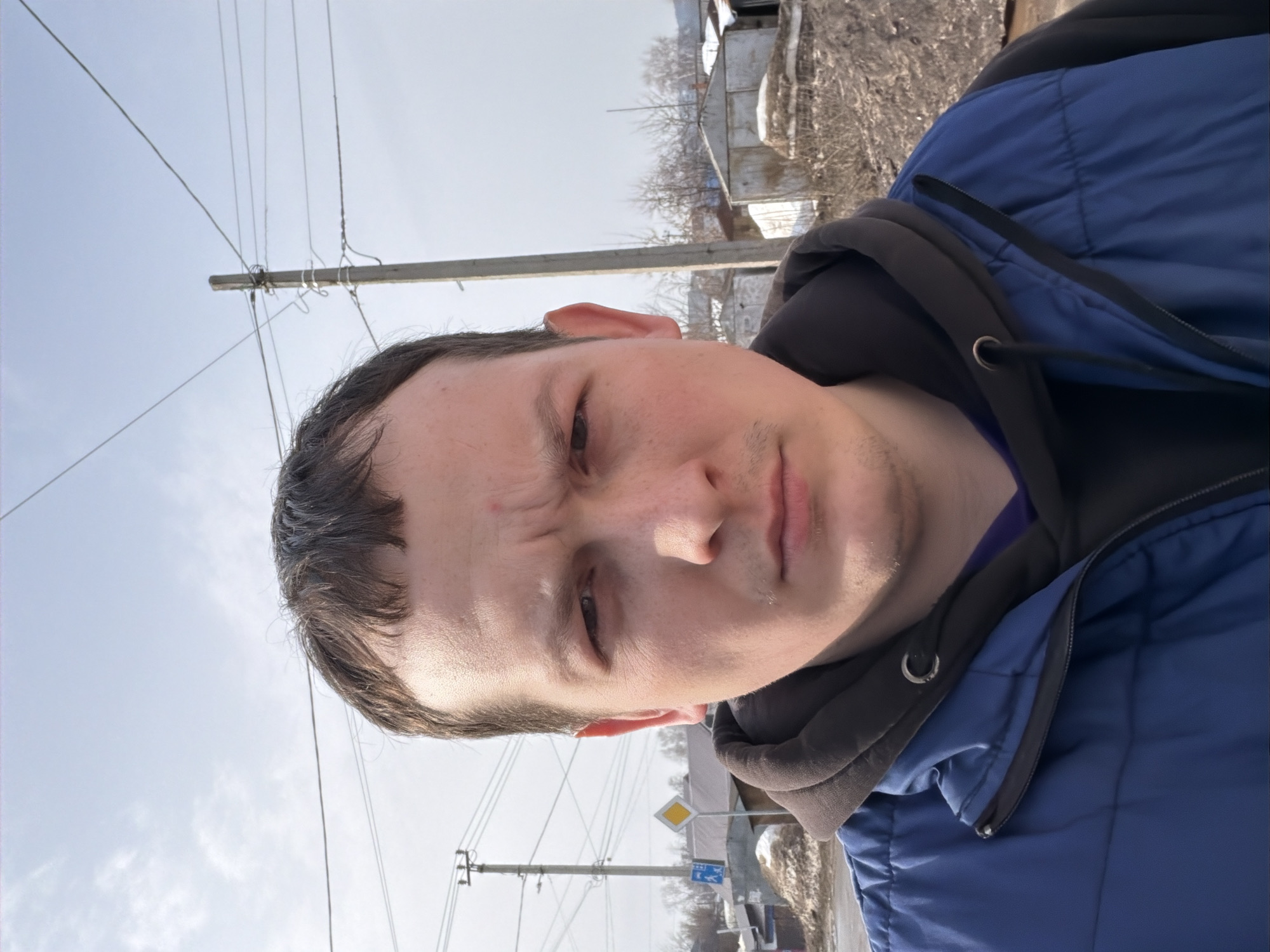 A male subject is depicted in a close-up, head-and-shoulders outdoor portrait. He has short, dark brown hair and minimal facial stubble, wearing a blue quilted vest over a dark brown hooded sweatshirt. His gaze is directed forward, exhibiting a slight furrow in his brow. The background features a partly cloudy light blue sky. To the subject's left, a yellow diamond-shaped road sign, indicating a priority road, is visible, with a smaller blue rectangular sign positioned beneath it. Multiple utility poles, including a prominent concrete pole on the right, support an extensive network of overhead power lines across the upper and middle ground. In the distance, several small, weathered, metal-clad structures, likely garages or sheds, are visible, along with sparse, bare trees and mounds of dirty snow or earth, indicative of a cold season. The environment is consistent with a semi-developed or industrial-residential area. While the image is stated to be from Chaikovsky, Russia, no specific identifying landmarks are present within the frame to allow for precise street-level geolocation within the city.