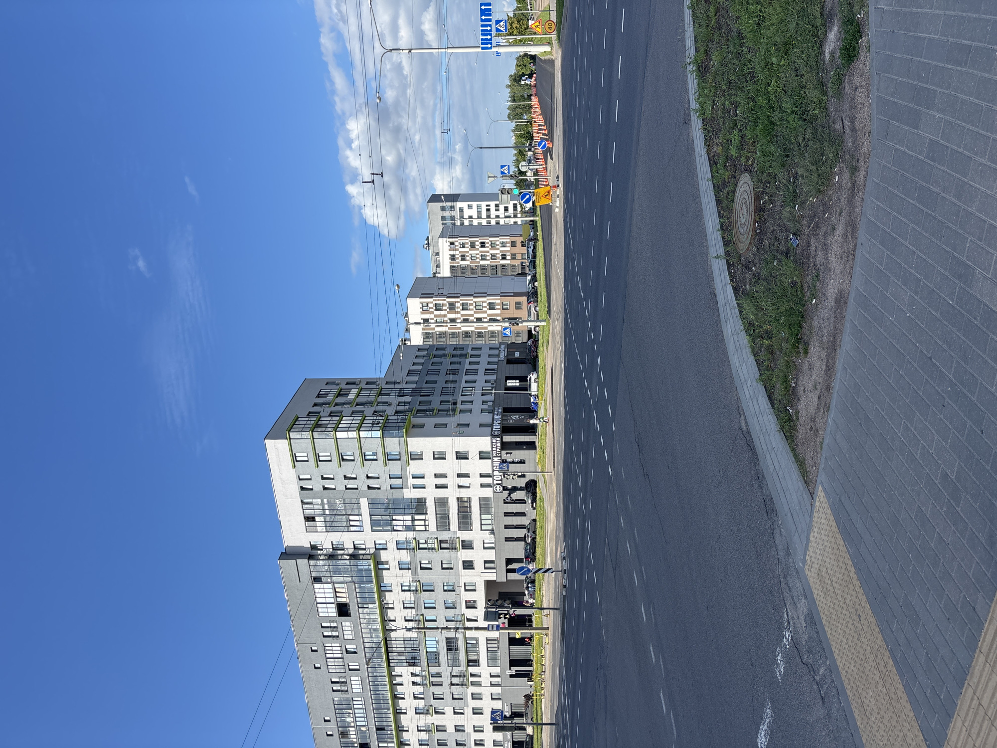 The image shows a street scene in Minsk, Belarus, on a clear day with scattered clouds. In the foreground, a paved road with lane markings and a pedestrian crossing is visible. To the right of the road, there is a sidewalk and a patch of grass with a manhole cover. In the background, several modern multi-story apartment buildings line the street. The buildings have a gray facade with white and green accents. Some of the buildings have large windows and balconies. Traffic signals, streetlights, and road signs are also present. A line of orange traffic cones indicates construction or road work further down the street. A few cars are parked along the sidewalk in front of the buildings, and a person is walking on the sidewalk. The overall impression is of a contemporary urban environment.