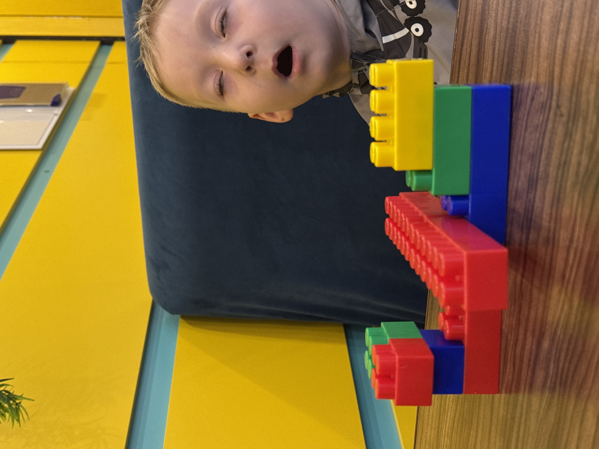 This image, likely captured indoors in Perm, Russia, is rotated 90 degrees clockwise, presenting a unique perspective. The central subject is a young child, possibly a toddler, whose fair-haired head and upper torso are visible from a high angle. The child has an open mouth, as if caught in a yawn, a surprised expression, or mid-vocalization, with eyes partially closed.
To the right of the child, a vibrant structure of large, primary-colored building blocks dominates the foreground. These construction blocks are yellow, green, blue, and red, stacked together. The structure appears to be built on or leaning against a wooden surface, which makes up the right side of the frame.
In the background, a dark blue upholstered surface, likely part of a chair or sofa, provides a soft contrast to the bright colors. Below this, a bright yellow floor or wall is visible, segmented by narrow, light blue strips. In the bottom-left corner, a small amount of green foliage can be seen. Another detail includes a white rectangular object with a dark square on top, resting on the yellow surface in the mid-left. No discernible text is visible on any objects in the image. The lighting appears bright and even, consistent with an indoor setting during daytime. The overall scene suggests a moment of playful interaction or observation.