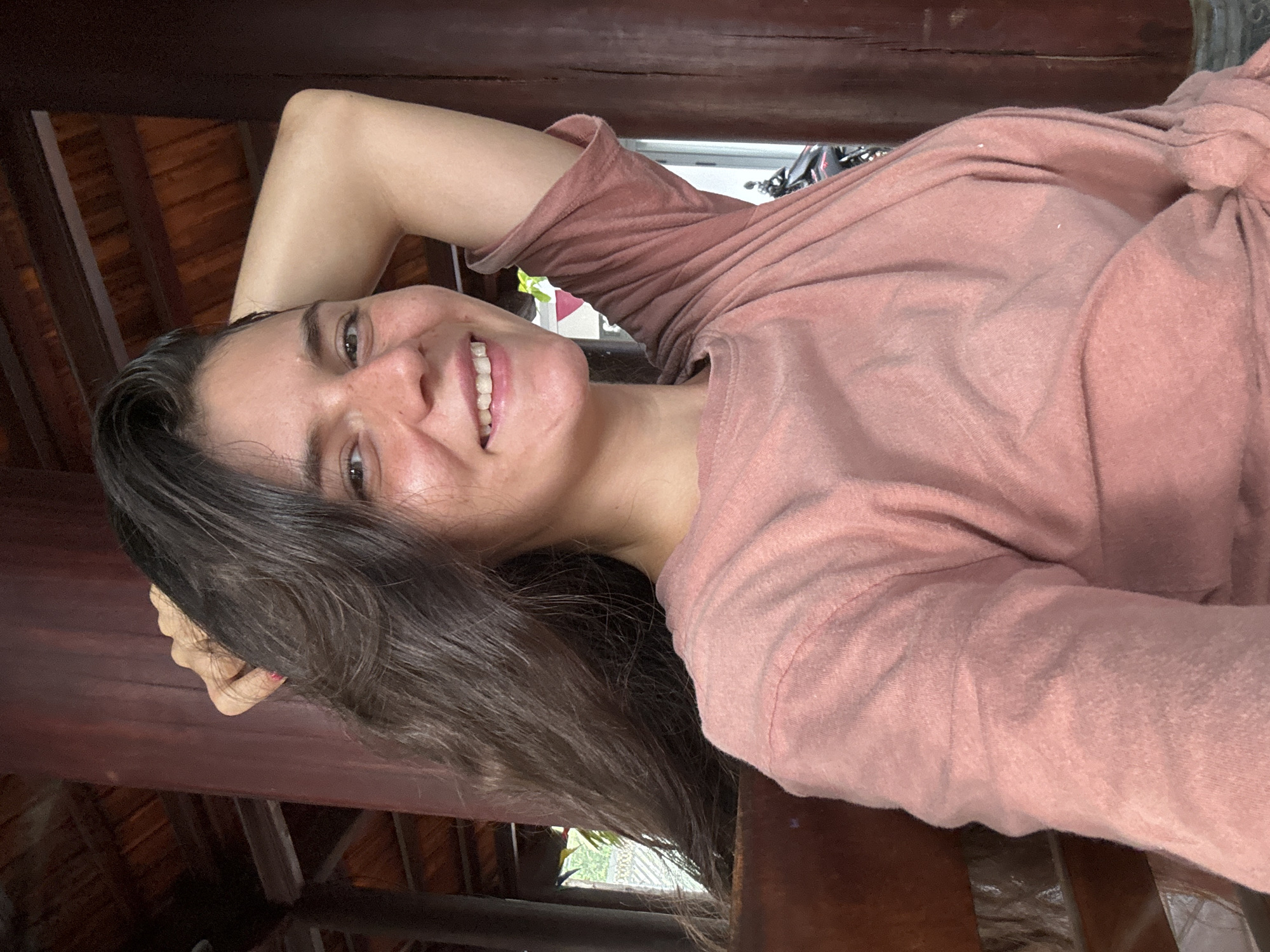 A young woman with long, dark, wavy hair is pictured from the chest up, sitting outdoors in what appears to be an open-air structure with dark wooden pillars and a wooden ceiling. She is looking towards the camera and smiling, with her right arm raised and bent behind her head, her hand resting in her hair. She is wearing a loose-fitting, muted pink t-shirt that is knotted on the left side. The background shows more of the wooden structure, with glimpses of greenery and a sliver of a dark motorcycle visible to the right. The overall lighting is soft and natural. The location is Mũi Né, Vietnam.