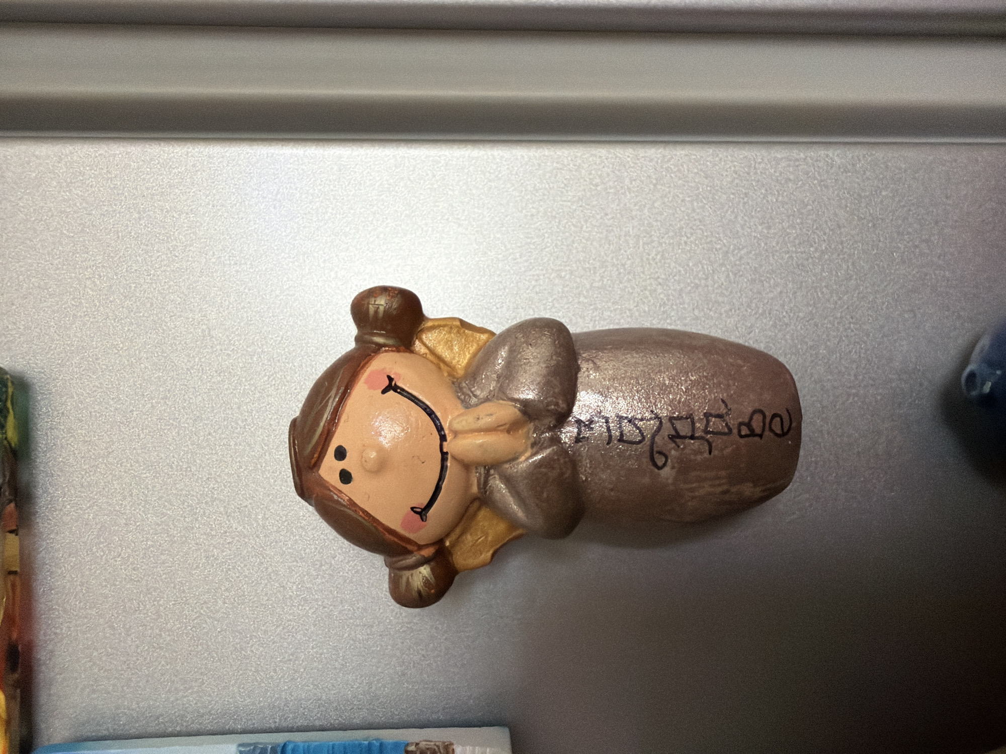 The image shows a close-up of a ceramic magnet shaped like a young girl with angel wings. The magnet is attached to a silver, textured surface, likely a refrigerator door. The girl has brown hair styled in two pigtails, a smiling face with rosy cheeks, and her hands are clasped together in front of her chest as if in prayer. The magnet is predominantly brown and gold, with black detailing for the eyes and mouth. There is black text written vertically down the front of the magnet, appearing to be in Hebrew script. The background is a plain, metallic surface with a vertical seam on the right side, indicating a large appliance. There are no people or other significant objects visible in the frame. The image does not contain any information that would allow for its location to be identified in the city of Pazardzhik, Bulgaria.
