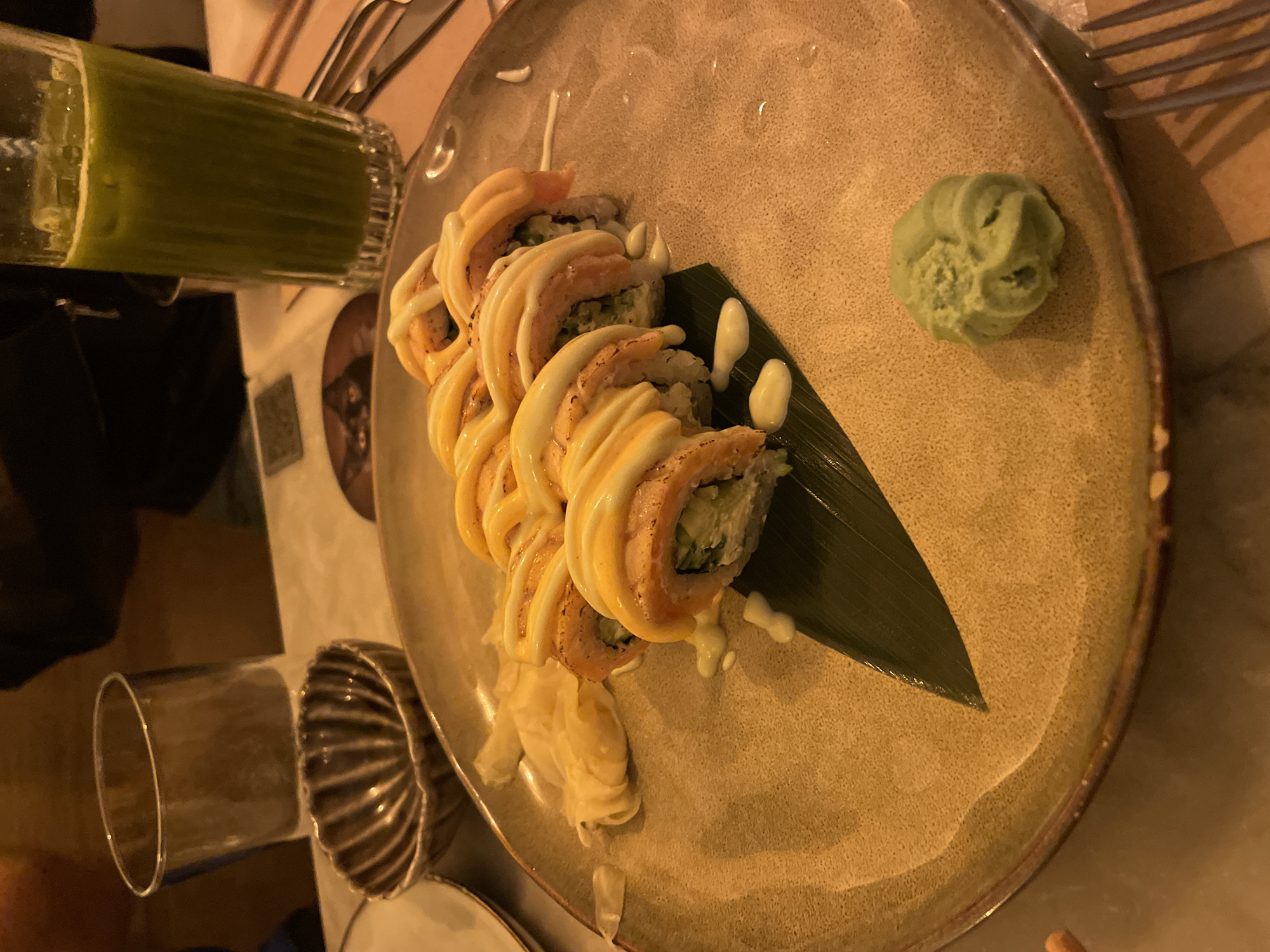 The image shows a plate of sushi, likely taken in an indoor restaurant setting. The plate is a light brown color, with a row of sushi rolls topped with a creamy sauce. There is a small mound of wasabi on the plate, and a few pieces of ginger. A green-colored beverage in a glass and silverware are visible on the table next to the plate. The setting is likely a restaurant in Barcelona, Spain.