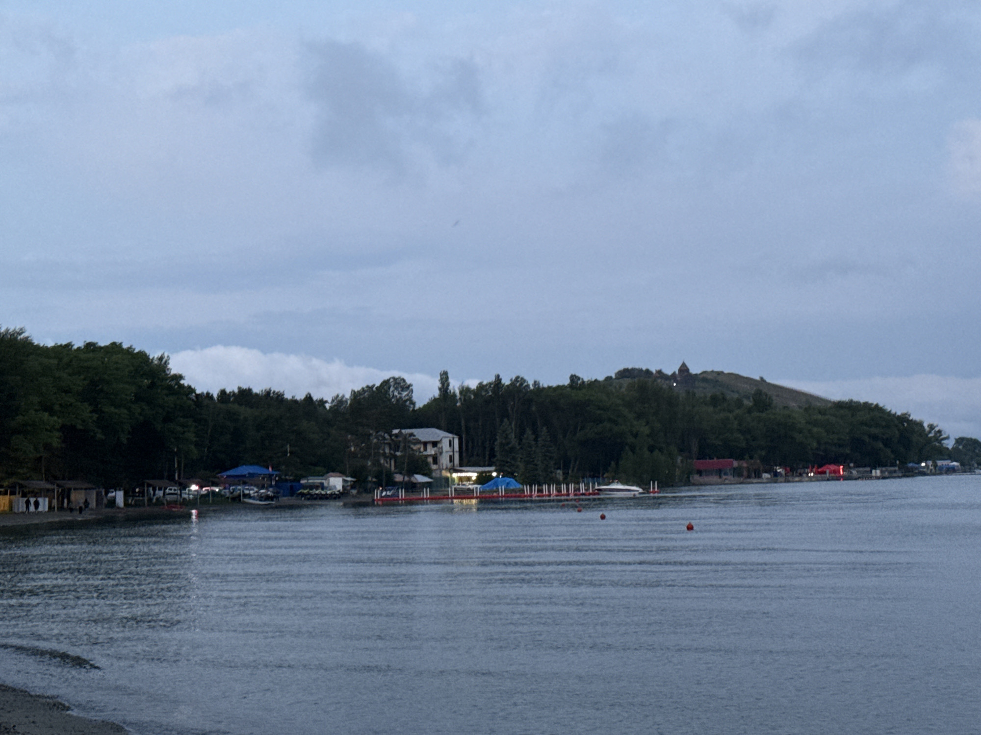 The image depicts a waterfront scene in Sevan, Armenia, likely taken during twilight or early evening. In the foreground, a body of water, presumably Lake Sevan, is visible, with gentle ripples on its surface and a faint reflection of lights from the shore. The water appears calm.
The middle ground features a developed shoreline with a mixture of structures and natural elements. Several small buildings, possibly kiosks or temporary shelters, line the beach area, with some people visible standing near them. A road or pathway runs behind these structures, with parked cars. Further along the shore, more substantial buildings are present, including a white multi-story building and structures with red and blue awnings, suggesting commercial establishments or recreational facilities. Several boats are docked or moored near piers and floating platforms. Red buoys are also scattered in the water.
The background is dominated by a dense line of trees that form a lush green backdrop to the shoreline. Beyond the trees, on a hill to the right, a distinctive architectural structure, possibly a church or historical monument, is visible against the dusky sky. The sky itself is overcast with diffused light, indicating cloudy conditions. The overall impression is one of a tranquil evening by the lake, with signs of human activity and a hint of historical presence on the hill.