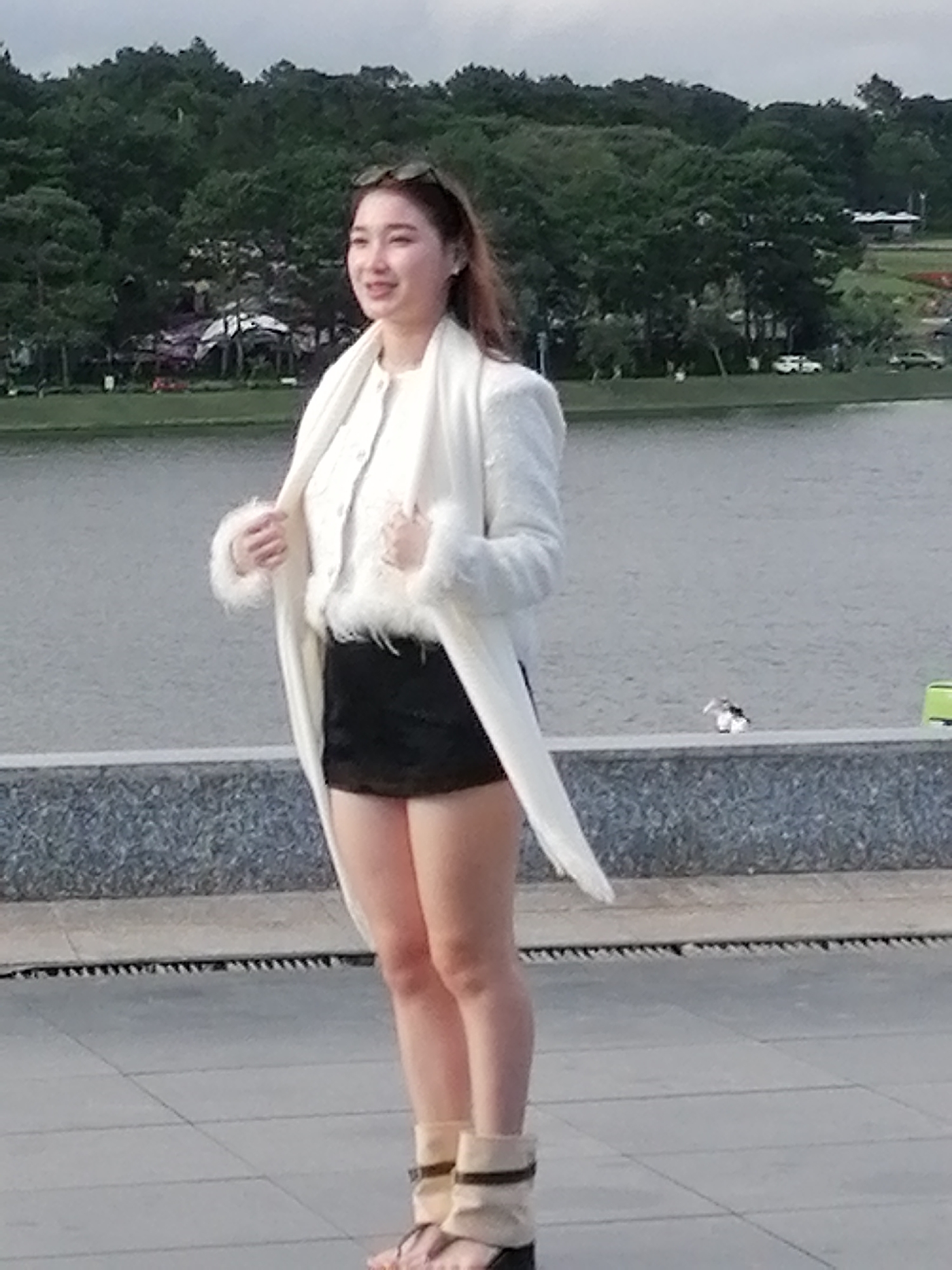 A woman stands on a paved walkway next to a body of water, with a tree-covered hillside in the background. She is wearing a white, fur-trimmed jacket over a white top and a black mini-skirt. She has on tall, beige sandals with dark straps. Sunglasses are perched on her head. The scene appears to be located at Xuan Huong Lake in Da Lat, Vietnam, based on the characteristic landscape.