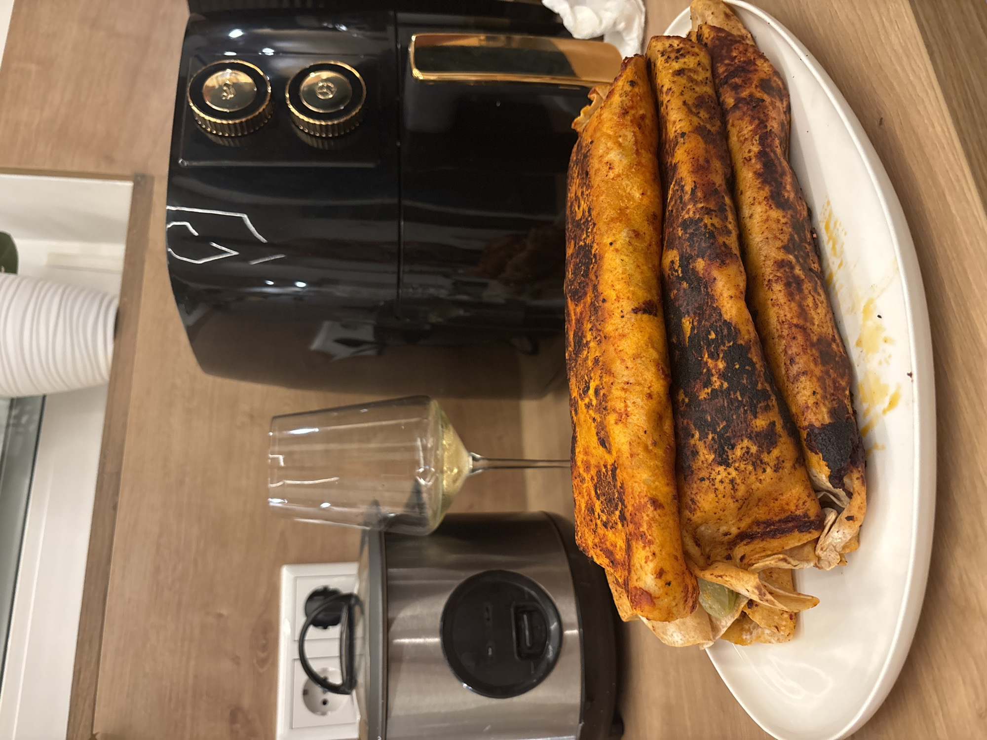 A stack of four golden-brown, slightly charred taquitos is presented on a white plate. The taquitos, rolled and filled, have a vibrant orange hue with darker, crispy edges. To the left of the plate sits a clear wine glass, partially filled with a pale yellow liquid, likely white wine. Behind the glass, a silver rice cooker with a black lid and control panel is visible. Further to the right, a prominent black air fryer with gold accents and two round knobs stands against a wooden background. A white electrical outlet with a black plug is also partially visible to the left of the rice cooker. The setting appears to be a kitchen counter in Viernheim, Germany.