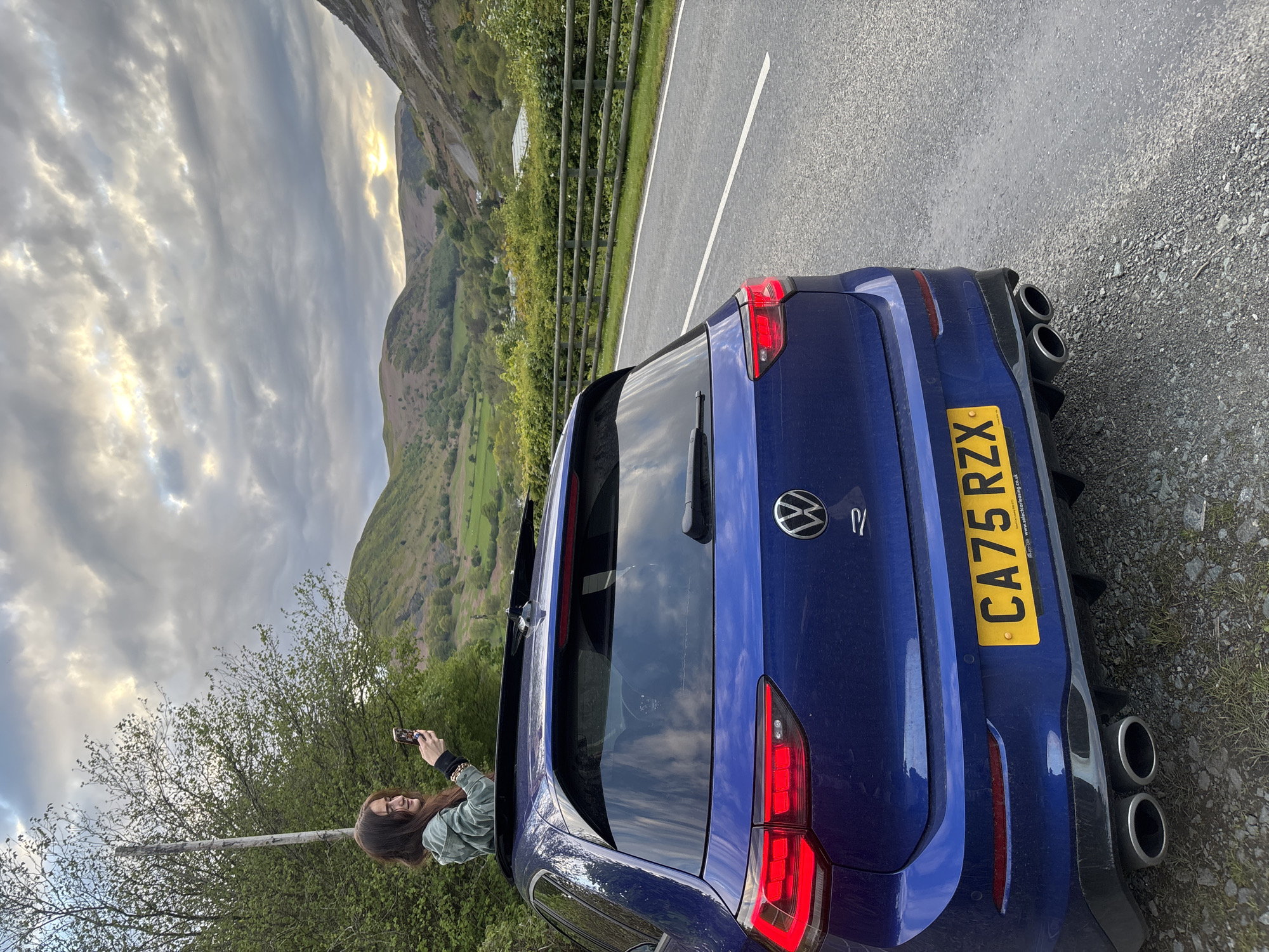 A blue Volkswagen Golf R with license plate CA75 RZX is parked on the side of a road. The vehicle is situated near a metal guardrail separating the road from a grassy hillside. A woman is seen standing near the car, holding a smartphone, possibly taking a photo. The surrounding environment includes rolling hills and mountains under a cloudy sky. The location is Llangynog, United Kingdom.