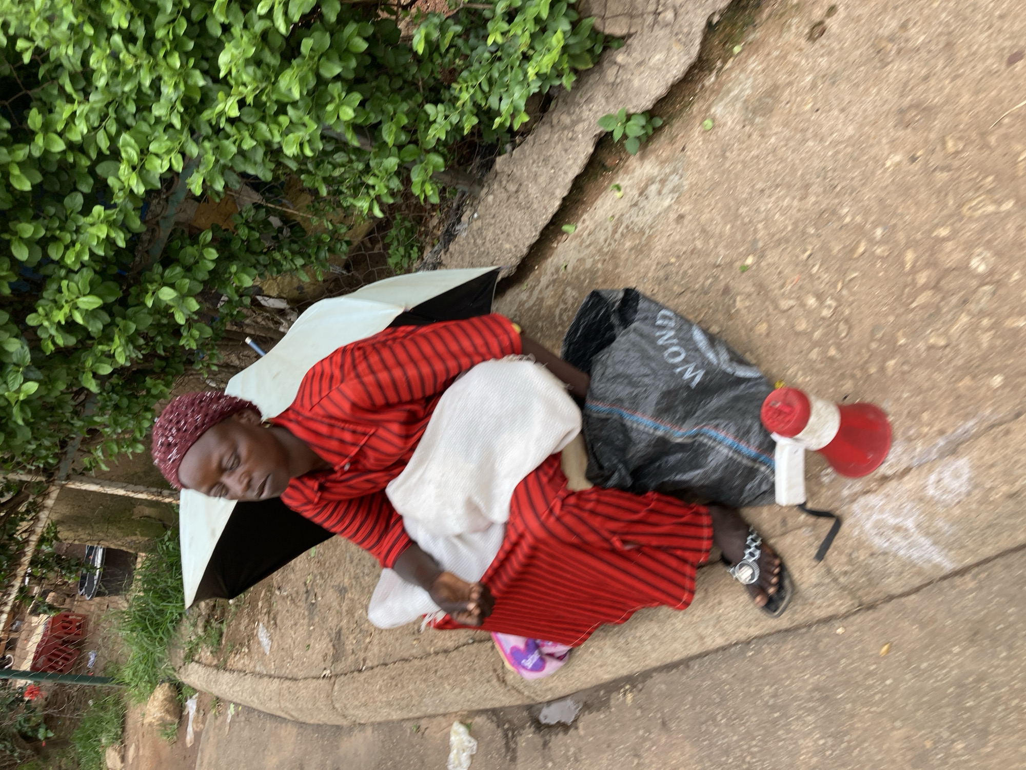 A person is sleeping on a concrete sidewalk, lying on their side under an open black and white umbrella. They are wearing a red and black striped shirt, a red head covering, and black sandals with a silver watch on their left ankle. White fabric is draped over their legs and torso. Next to them is a black plastic bag with white lettering that appears to spell "WONDER" and a red and white megaphone. The scene is outdoors, with lush green foliage visible to the left of the sidewalk and a chain-link fence behind it. The ground is concrete, with some cracks and debris. The overall impression is one of homelessness or extreme poverty, with the individual seeking shelter from the elements on the street. The lighting suggests it is daytime, likely with overcast or dappled sunlight, given the lack of harsh shadows. There are no strong cues about the specific time of day or weather conditions beyond the use of an umbrella. The location context is Jos, Nigeria.