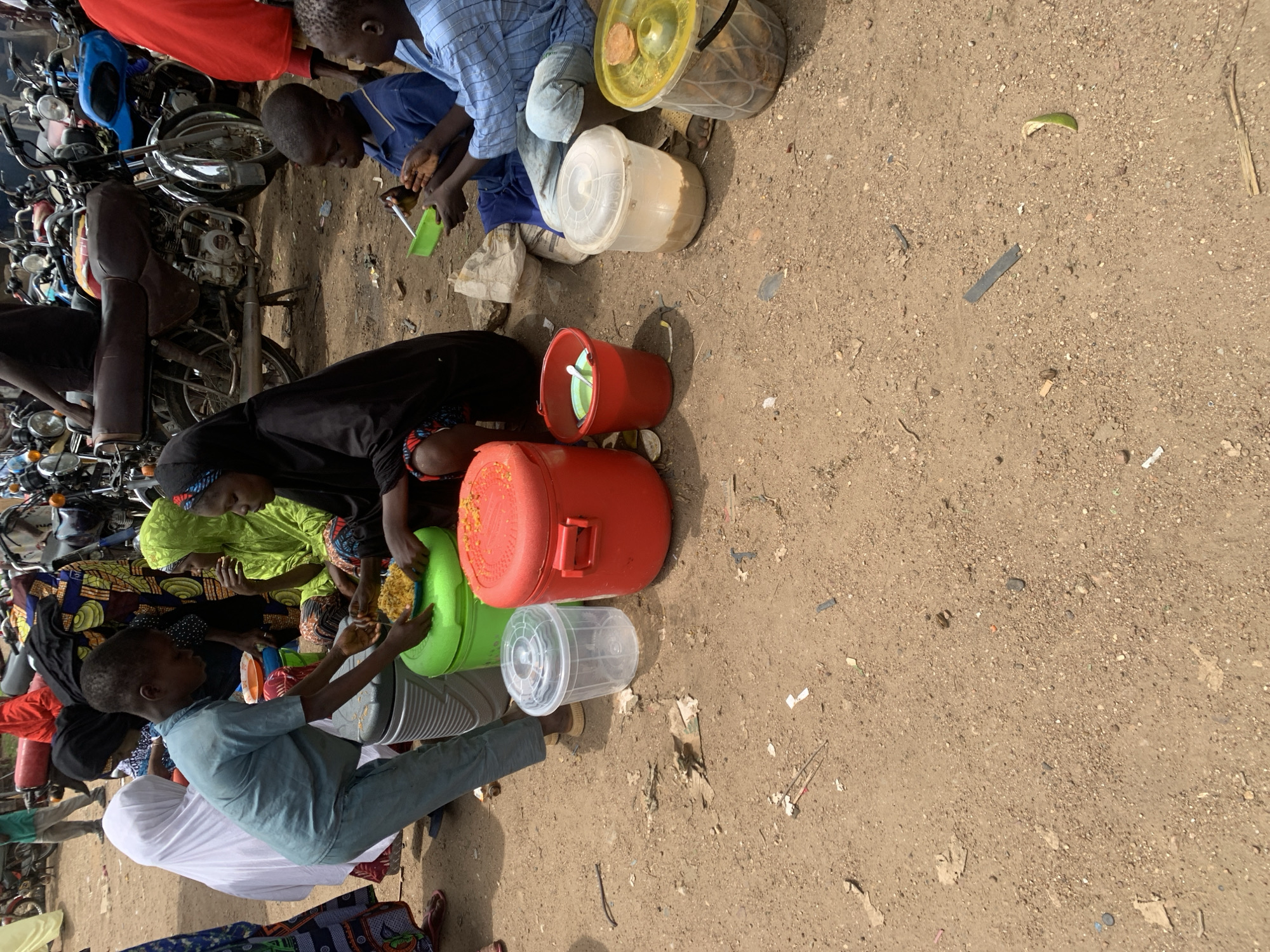 In Project, Nigeria, a group of children and adults are gathered outdoors on a dirt ground, possibly at a market or communal gathering. In the foreground, a young boy in a blue shirt and pants is serving food from a large red container to a child wearing a blue shirt. To the left, a girl in a white hijab stands with a large green container. Several other people in traditional attire are seated on the ground, including a woman in a green headscarf and a woman in a black headscarf. Various containers, including large plastic tubs and buckets, are scattered around the scene. In the background, numerous motorcycles are parked, suggesting a busy area. The environment appears somewhat dusty and unpaved, typical of many areas in Nigeria.
