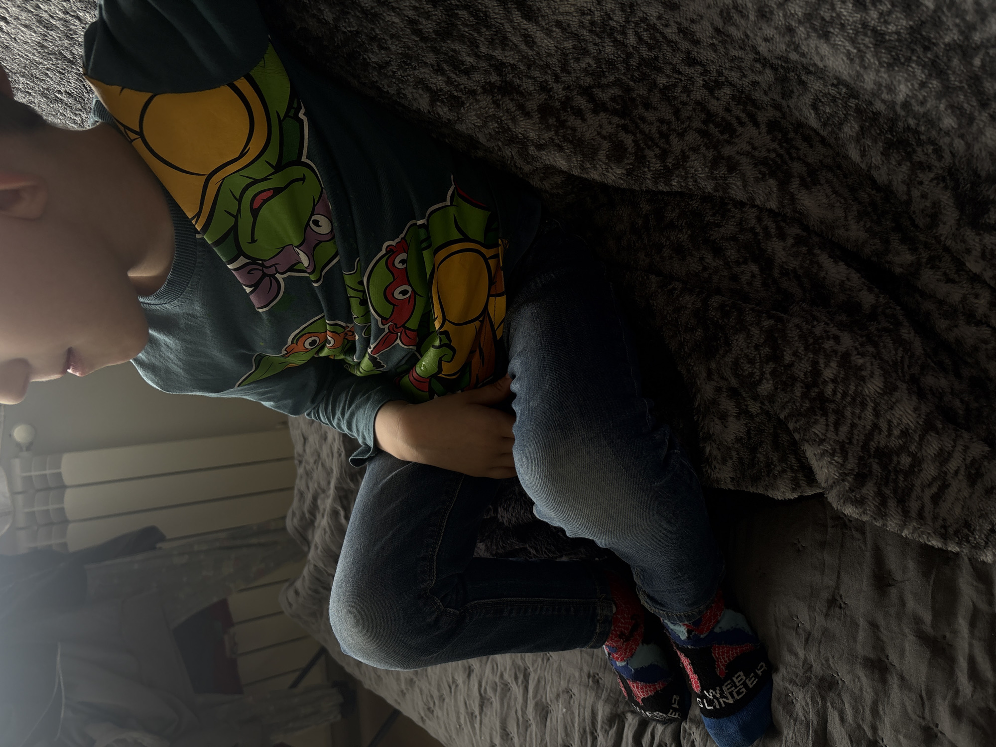A boy lies on a bed in an interior location. He is wearing a Teenage Mutant Ninja Turtles shirt, blue jeans, and blue socks that say "Web Slinger". The bed has a gray duvet. Part of a radiator and a dim reflection of the room are visible in the lower left corner. The location appears to be indoors, but the image does not provide enough information to determine if it is in Sofia, Bulgaria.