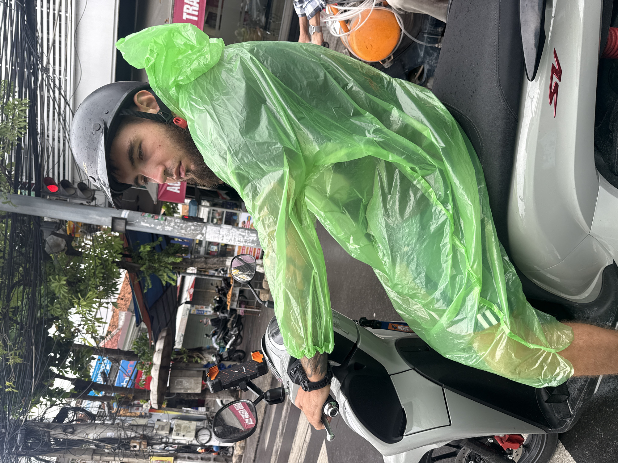 An adult male wearing a black helmet and a bright green disposable raincoat is seated on a white scooter on a street in Nha Trang, Vietnam. The man has a beard and is looking to his right, away from the camera. His right hand is on the scooter's handlebar. The scooter's seat is black and appears wet. In the background, there are shops, trees, power lines, and other motorcycles. The scene suggests it is raining, as indicated by the wet surfaces and the man's raincoat. The image is captured from a slightly elevated angle, focusing on the man and the scooter.