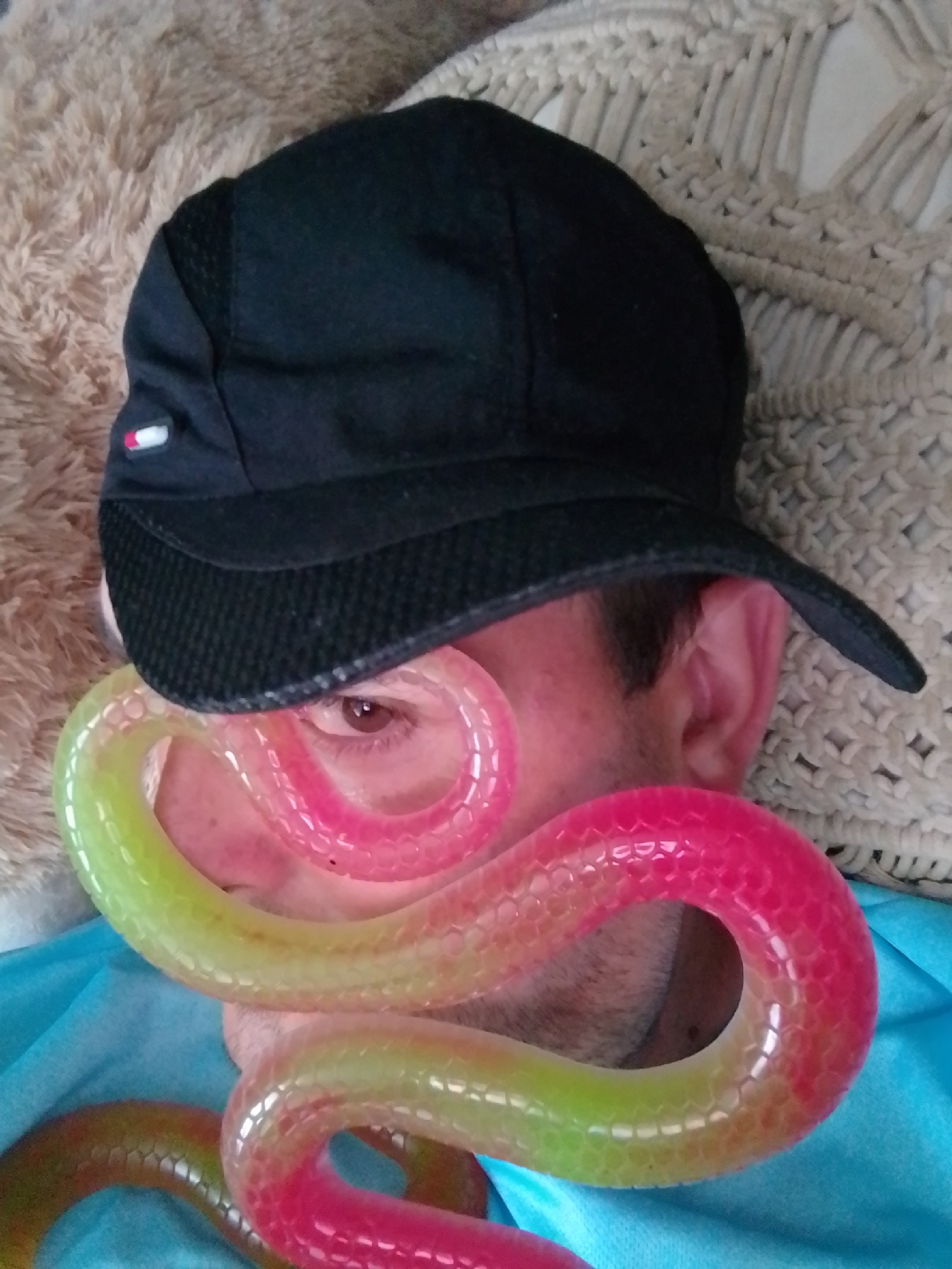 A person wearing a black baseball cap and a blue shirt is lying down. A colorful, segmented tube, possibly a toy, is coiled around the person's face, obscuring their nose and mouth. The tube transitions in color from green to pink. The person's eye is visible. The background consists of a textured, cream-colored fabric and a fluffy, beige material. The image does not provide any information about a specific location in the city of Wedel, Germany.