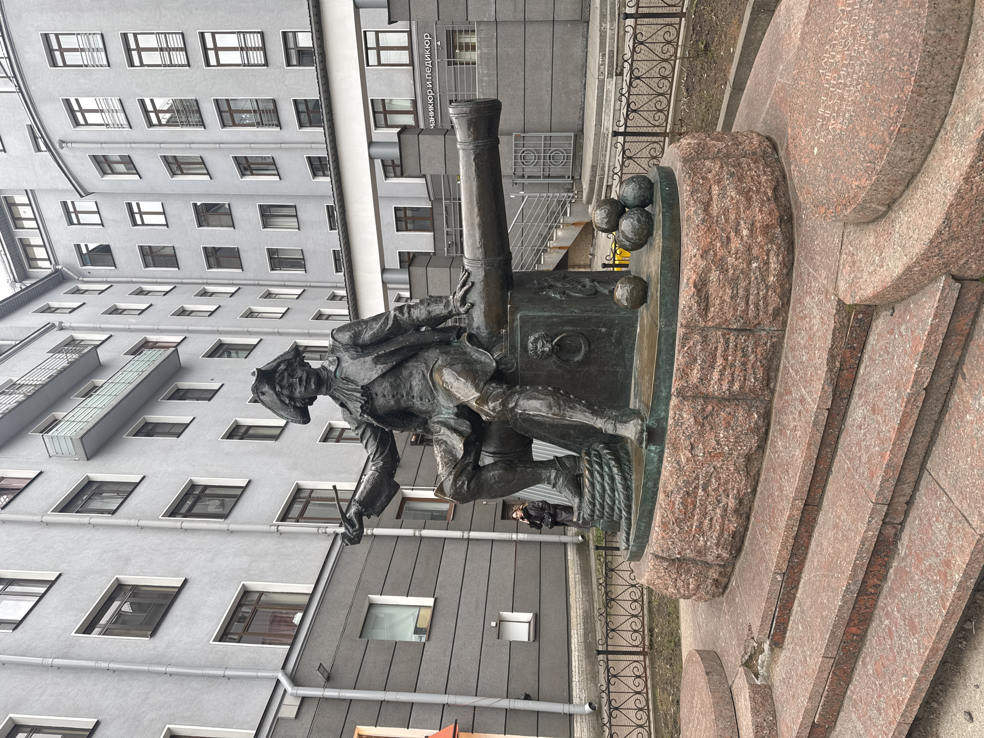 The image displays a bronze monument featuring a historical figure and a cannon, set against a modern urban backdrop. The central subject is a bronze statue of a man, likely Peter the Great, dressed in period attire including a tricorne hat, a long coat, and boots. He holds a ramrod or similar implement, appearing to prepare the adjacent large bronze cannon. Three bronze cannonballs rest on the monument's base next to the cannon.
The monument stands on a multi-tiered pedestal constructed from reddish-brown granite. In the background, a tall, grey-facaded building with numerous rectangular windows is prominent. The building has a modern, somewhat industrial appearance with visible drainage pipes and some windows featuring small balconies or security bars. A decorative black metal fence runs along the right side of the scene. A person in dark clothing is partially visible standing on the pavement in the mid-ground, near the monument's base, providing a sense of scale.
The scene is captured during the daytime under an overcast sky, indicated by the soft, diffused light and lack of strong shadows. On the building in the background, Cyrillic text "МАНИКЮР И ПЕДИКЮР" (MANICURE AND PEDICURE) is visible, indicating a business. Faint, engraved Cyrillic text is also present on the granite base of the monument, including "САНКТ-ПЕТЕРБУРГ" (SAINT PETERSBURG) and other historical references such as "АВГУСТА МЕСЯЦА" (month of August).