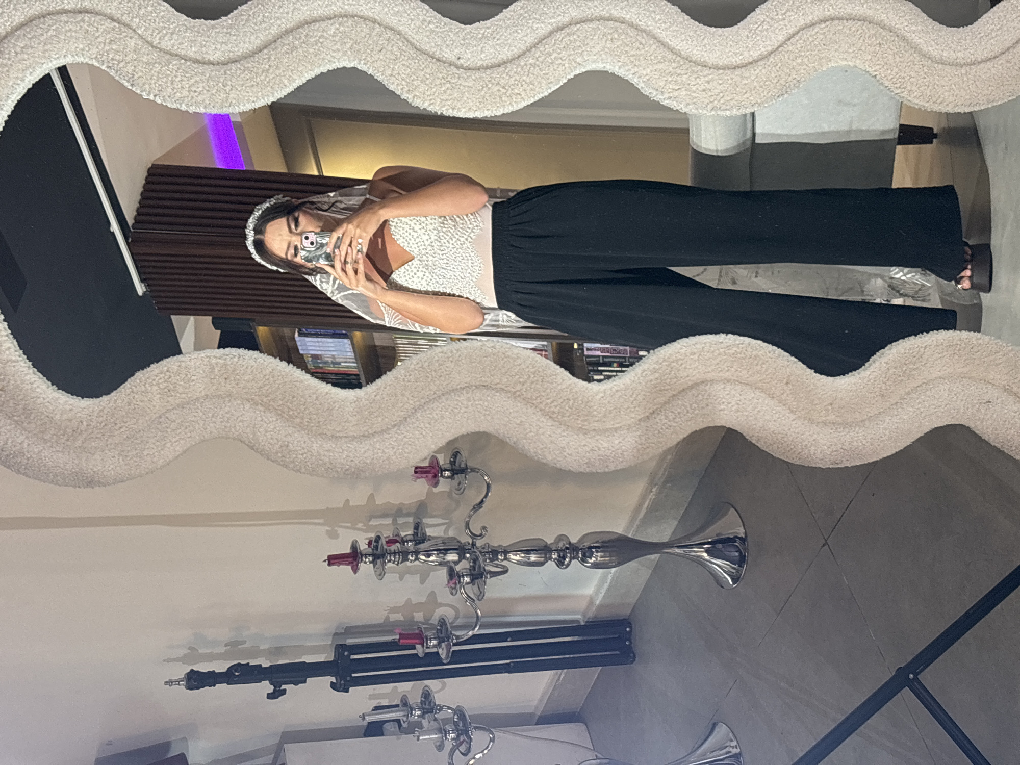 The image is a vertically oriented, full-body mirror selfie taken in an indoor setting, likely a home. The subject, a woman, is wearing a white, sequined top with a sheer overlay and black, wide-leg pants. She has on a sparkling headband and is holding a smartphone in her hands, capturing the reflection. The mirror itself has a textured, fluffy white border with a wavy pattern.
In the reflection, we see the woman from the chest up, holding her phone to take the picture. She appears to be in a room with wooden paneling on the wall behind her, and a hint of purple light from what looks like a shelf or accent lighting. Below her, reflected in the mirror, is a sleek, silver candelabra with several pink candles. There are also reflections of other objects, including what appears to be part of a camera tripod and possibly some books on shelves in the background. The floor in the reflected view is tiled. The lighting suggests it is daytime or well-lit interior. The overall mood is casual and self-aware. The location context provided is Sweileh, Jordan.