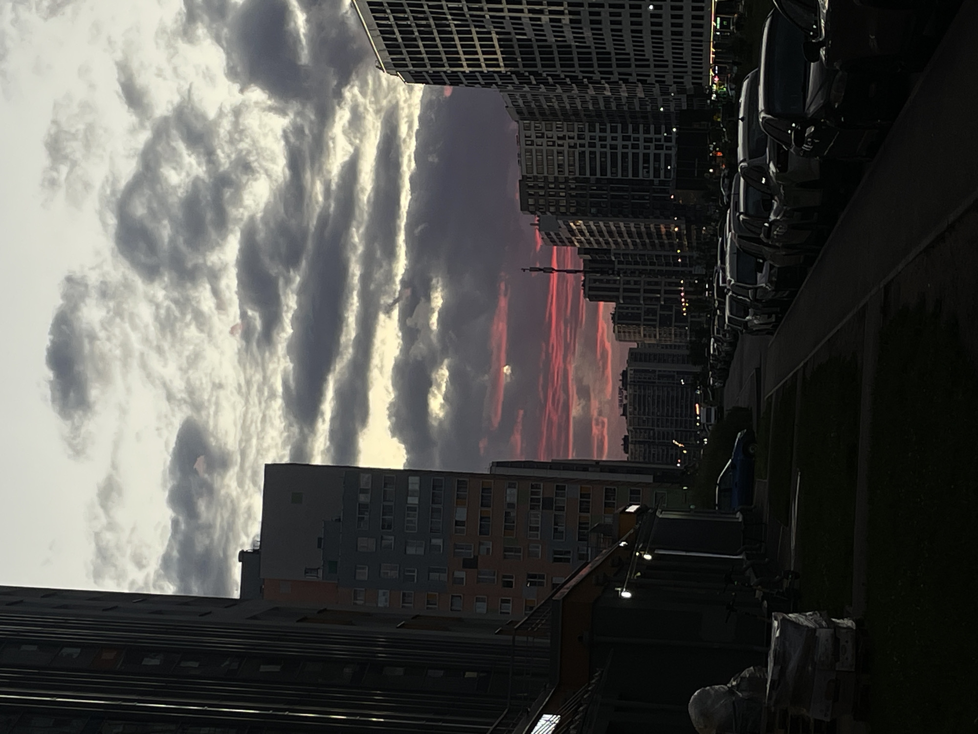 The image depicts a street scene in a modern urban environment, likely a residential area in Russia, as indicated by the architecture. The time of day appears to be dusk or early evening, with a dramatic sky filled with dark, cloudy formations tinged with pink and orange hues from the setting sun. A small, pale circle, possibly the moon or a planet, is visible within the colorful sky.
Dominating the left side of the frame are tall, multi-story buildings with dark facades and numerous windows. To the right, a row of similarly modern, high-rise apartment buildings stretches into the distance, their windows illuminated by interior lights, creating a grid-like pattern. Between these buildings, a street is lined with parked cars, their forms silhouetted against the fading light.
In the foreground, on the left, a dark building with storefronts is partially visible. Some external lights are on, illuminating the ground level. To the right of this building, a patch of dark grass and a sidewalk are present. A blue car is parked on the street further down, and more cars are visible parked along the right side of the street, forming a long line. In the lower left corner, stacks of construction materials or supplies are visible, covered in plastic sheeting. No people are visible in the scene. The overall impression is one of a contemporary city transitioning from day to night, with a striking sky as a focal point.